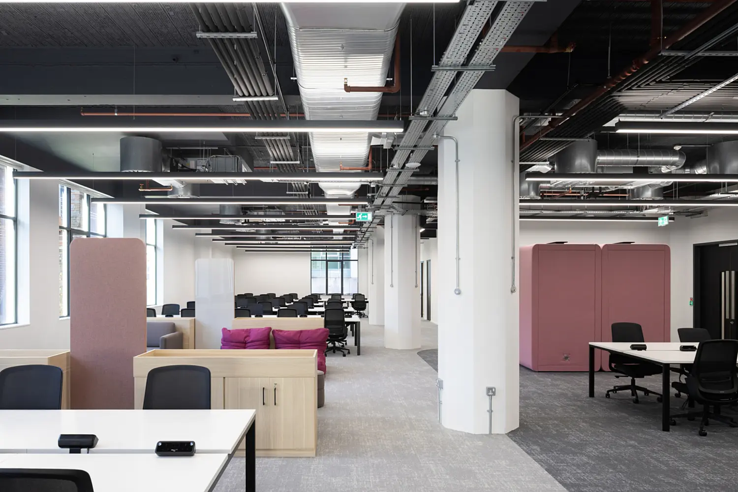 Modern open plan office workspace with a contemporary fit out and exposed ceiling featuring ductwork, cable trays and linear LED lighting arranged in parallel lines across the space. The layout is defined by large white structural columns that run through the centre, creating depth and subtle division between zones.  In the foreground, white desks with integrated power modules are paired with black task chairs, set on grey carpet tiles. To the left, a breakout seating area includes a light wood storage unit, a grey sofa and a cluster of bright pink lounge cushions positioned low to the floor, adding contrast to the neutral palette. A tall upholstered acoustic screen in soft pink stands nearby, helping to define the seating zone.  Further into the space, rows of desks extend towards the back, forming a structured working area with consistent black chairs and white surfaces. On the right, two large pink acoustic pods provide enclosed spaces for private calls or focused work, positioned near additional meeting tables and chairs.  Natural light enters from windows along the left side, while the combination of grey flooring, white walls and light wood finishes creates a clean, cohesive environment with bold pink accents throughout.
