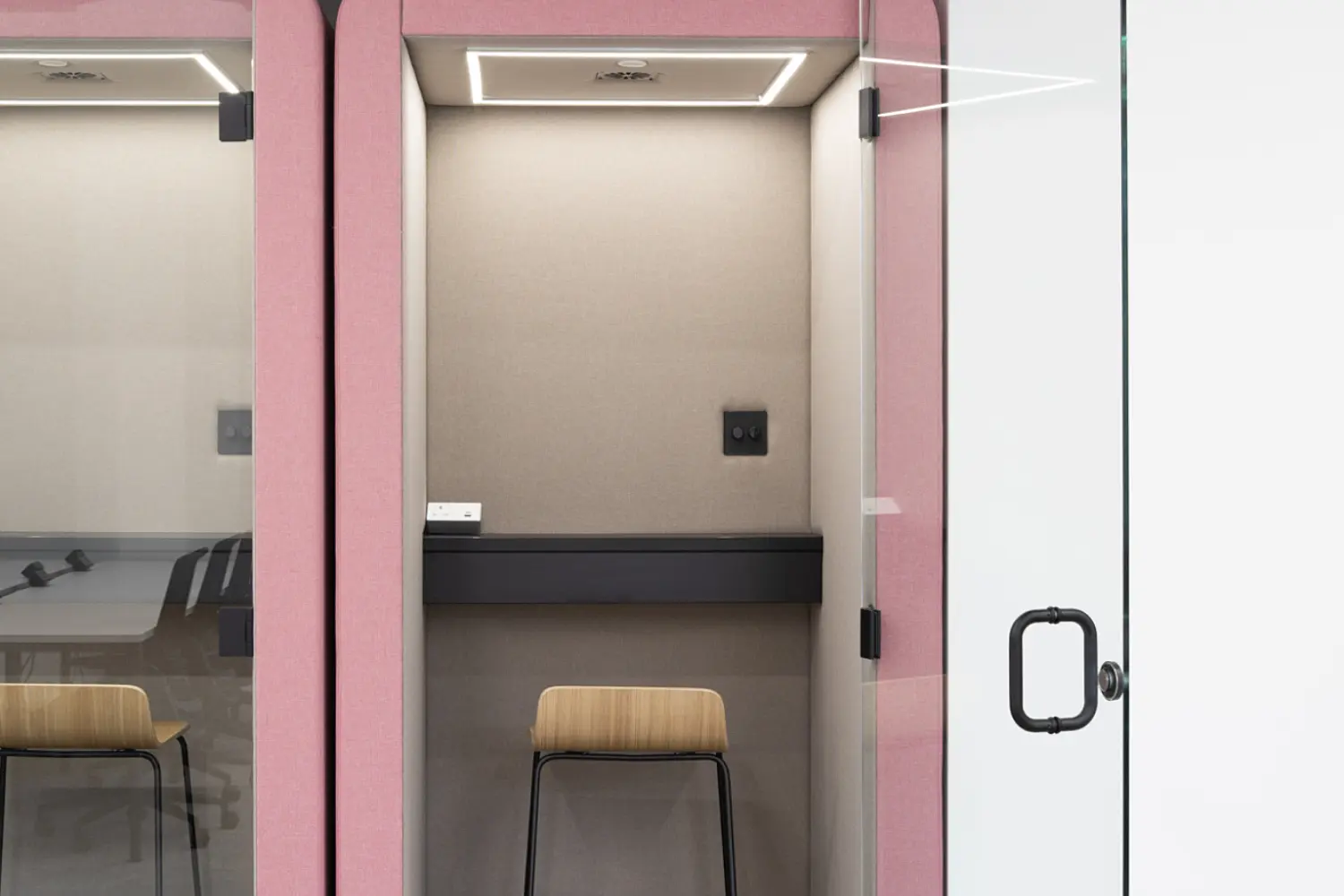 Modern office acoustic booths designed for private work or calls, finished in soft pink upholstered panels. Two adjoining pods are shown, with the right pod open and fitted with a clear glass door featuring a black handle.  Inside the open booth, a compact workspace includes a dark work surface, a high wooden stool with black legs and integrated power sockets on the back wall. A recessed ceiling light provides focused illumination within the booth, creating a quiet, enclosed environment.  The adjacent booth on the left is partially visible, also enclosed with glass and fitted with similar internal features. The surrounding space has a neutral finish with light grey flooring and white walls, allowing the pink booths to stand out as a bold yet functional element within the office fit out.