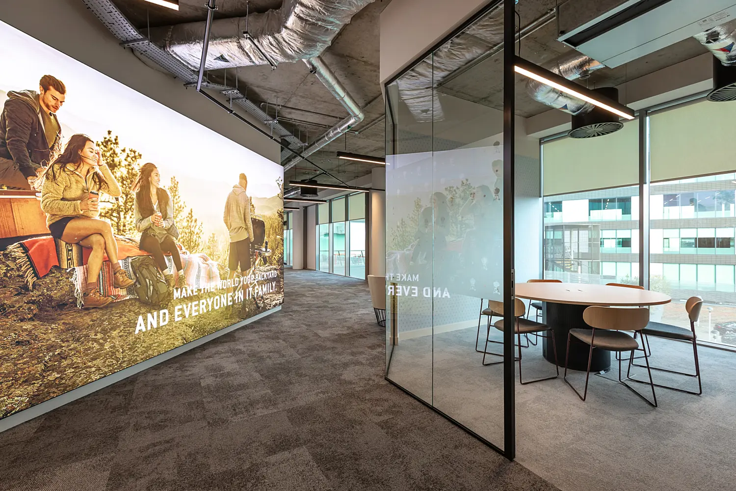 Office corridor with a contemporary design and exposed industrial ceiling featuring visible ductwork and linear lighting. On the left, a large wall graphic shows a group of people outdoors sitting on a vehicle, with visible text reading “MAKE THE WORLD YOUR BACKYARD AND EVERYONE IN IT FAMILY.” On the right, a glass meeting room with black framing contains a round table and several chairs, positioned beside floor to ceiling windows with roller blinds and views of neighbouring buildings. The corridor continues into the distance with additional glass fronted rooms, while the floor is finished in dark grey carpet tiles throughout.