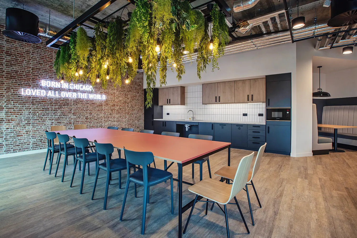 Modern office kitchen and dining space with an industrial style ceiling featuring exposed ductwork, pipes and suspended lighting. A large rectangular communal table with a muted red surface sits in the centre, surrounded by mixed chairs in blue, green and light wood finishes. Above the table, a black metal frame supports hanging greenery and warm exposed bulbs, creating a canopy effect.  Along the back wall, a kitchenette includes dark blue and wood effect cabinets, a white tiled backsplash and integrated appliances. To the left, a brick feature wall displays illuminated text reading “BORN IN CHICAGO LOVED ALL OVER THE WORLD.” On the right, a recessed booth seating area with upholstered benches and a small table sits within a softly lit alcove. The floor is finished in light wood effect planks, contrasting with the industrial ceiling above.