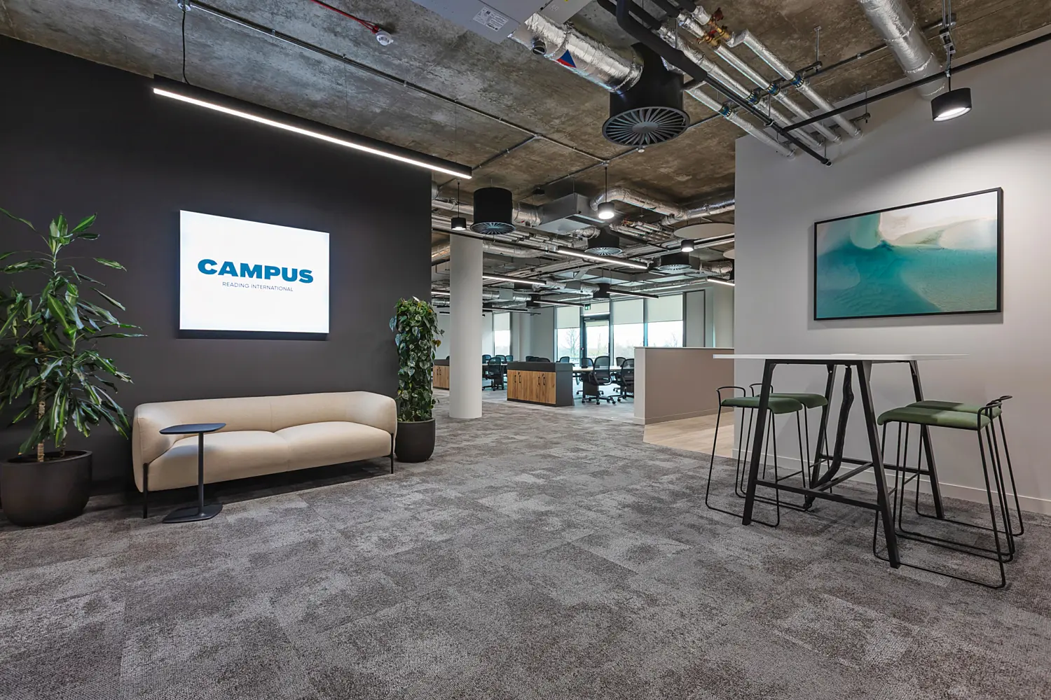 Modern office reception and breakout area with an industrial style ceiling featuring exposed ductwork, pipes and suspended linear lighting. On the left, a black feature wall displays a lit sign reading “CAMPUS READING INTERNATIONAL,” positioned above a curved beige sofa and a small round side table, with a tall potted plant nearby.  In the centre and background, an open plan workspace with bench desks and office chairs extends towards large windows, allowing natural light into the space. On the right, a high table with a white top is paired with tall stools featuring green upholstered seats, positioned beneath a framed artwork with abstract blue and green tones. The floor is finished in grey carpet tiles, tying the different zones together.