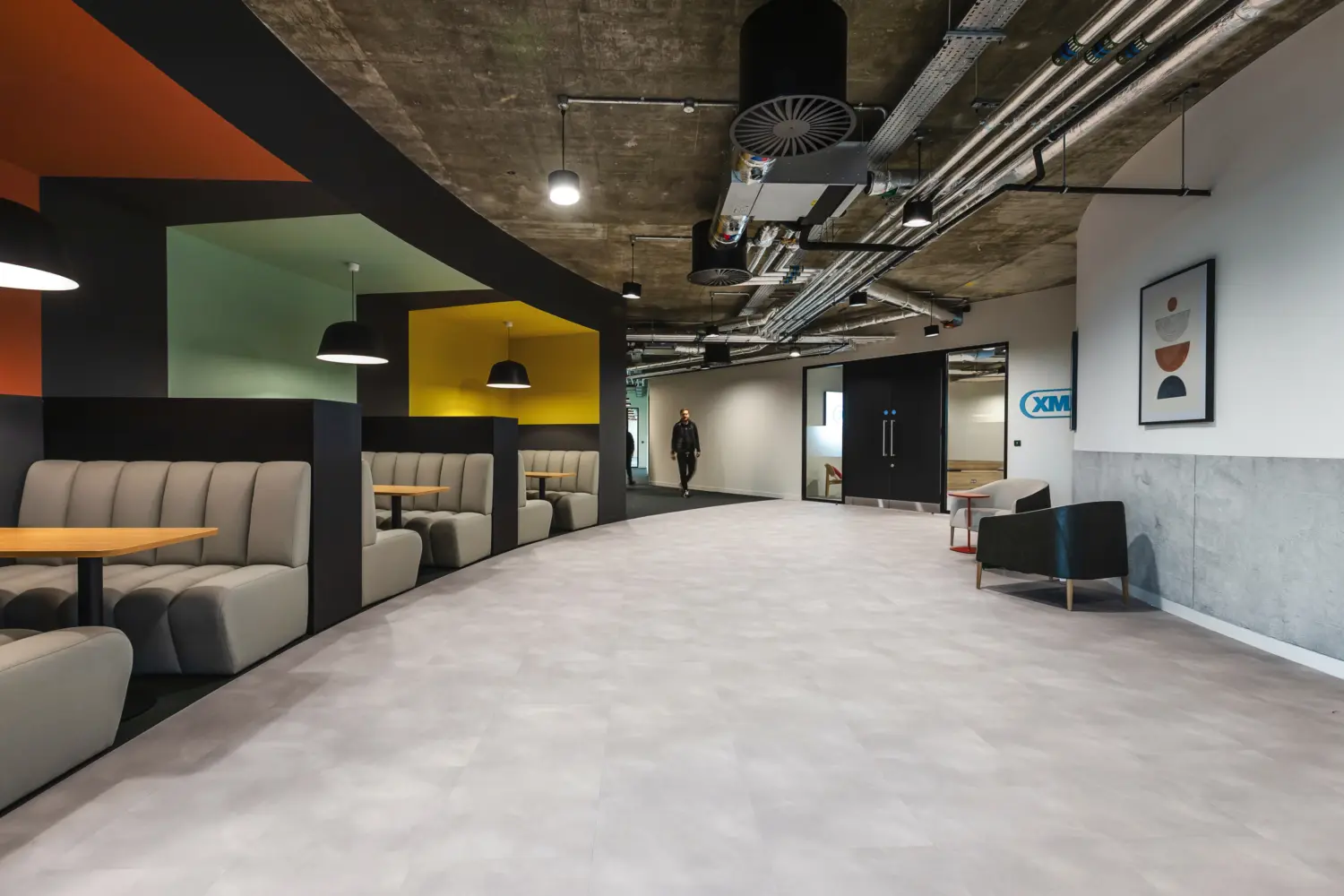 Modern office breakout area with an open industrial ceiling featuring exposed ductwork and lighting. On the left, a row of upholstered booth seating in neutral tones lines the wall, each booth paired with a small round wooden table and set within colourful alcoves in shades of green, yellow and orange. Pendant lights hang above the seating. The centre of the space has a wide, light grey floor leading towards a corridor, where one person is walking in the distance. On the right, there is a lounge area with a cushioned chair, a small table and framed abstract artwork on a white and grey wall.