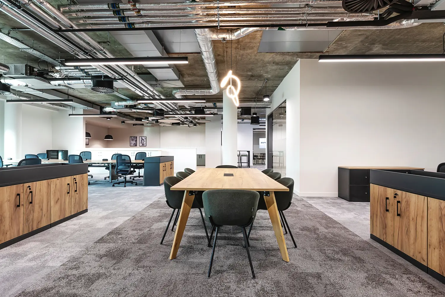 Open plan office workspace with a contemporary industrial aesthetic, featuring an exposed concrete ceiling with visible ductwork, pipework and suspended linear lighting. In the foreground, a large rectangular meeting table with a light wood surface is surrounded by dark green upholstered chairs. Above the table, a sculptural hanging light fixture with looping illuminated forms is suspended from the ceiling.  On either side of the space, low storage units with wood effect fronts and dark worktops provide separation between zones. In the background, bench desks with teal office chairs are arranged in an open working area, while to the right a glass partition encloses a meeting room. The floor is finished with grey carpet tiles throughout, creating a consistent base across the workspace.