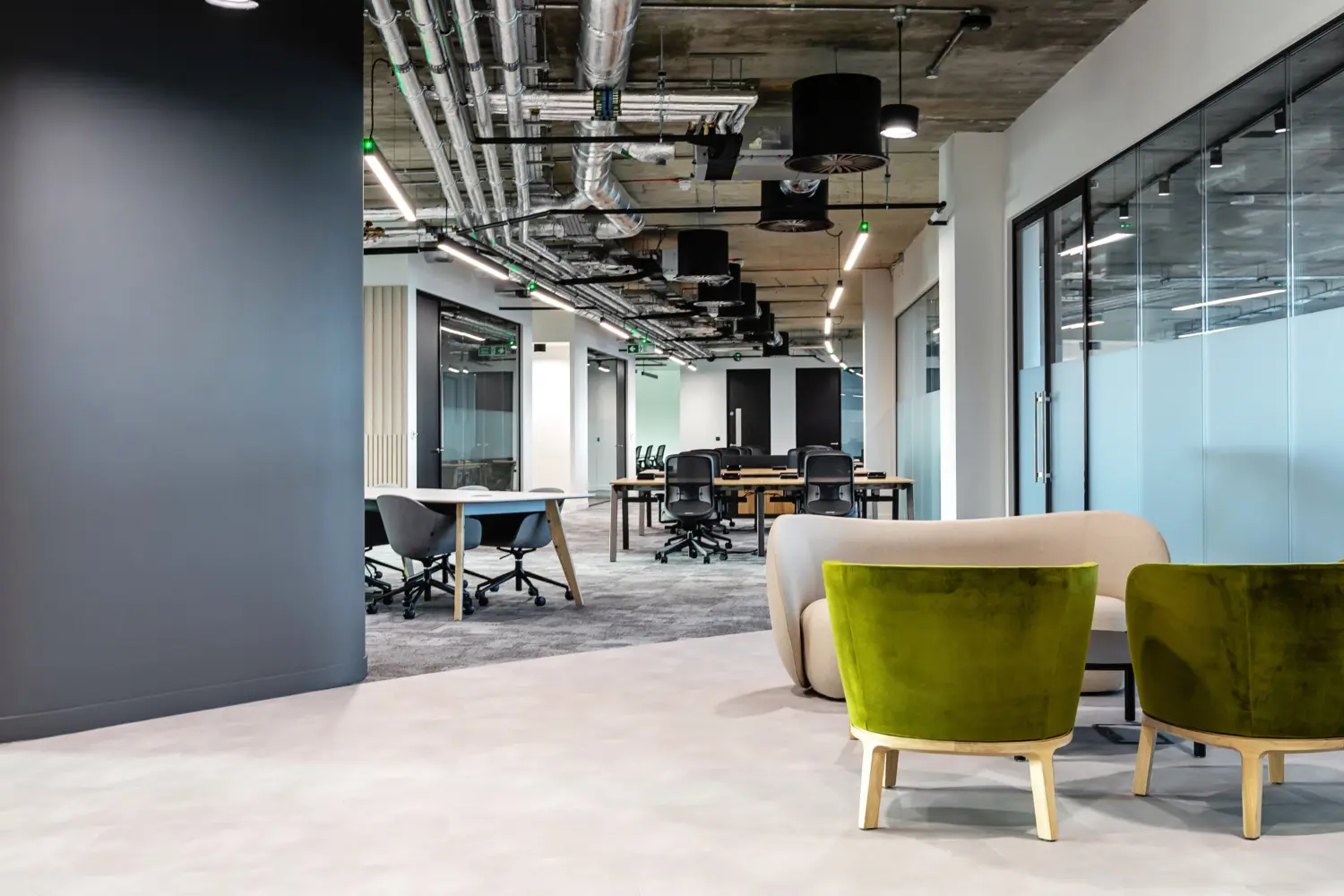 Modern open plan office area with an industrial ceiling featuring exposed ductwork, pipes and suspended linear lighting. In the foreground on the right, two green upholstered lounge chairs with light wood legs are positioned beside a curved beige sofa, forming a small seating area on a smooth, light grey floor.  Beyond this, the space transitions into a workspace with shared desks and black office chairs arranged in rows. To the right, a glass partition with a frosted lower section encloses meeting rooms, while to the left a dark curved wall creates a soft division between zones. The flooring shifts to grey carpet tiles in the main workspace, adding contrast between the lounge and desk areas.