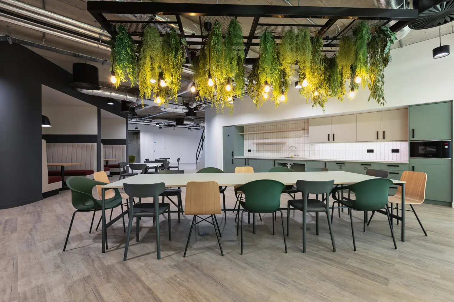 Modern office kitchen and dining area featuring a large communal table surrounded by mixed chairs in green and light wood finishes. Above the table, a suspended black metal ceiling frame holds dense hanging greenery and exposed warm light bulbs, creating a canopy effect. The space has an industrial ceiling with visible ductwork and lighting. At the back, a compact kitchenette with pale green and beige cabinets, a white tiled backsplash and integrated appliances runs along the wall. To the left, additional booth seating in neutral tones sits within a darker alcove, while the floor throughout is finished with light wood-effect flooring.