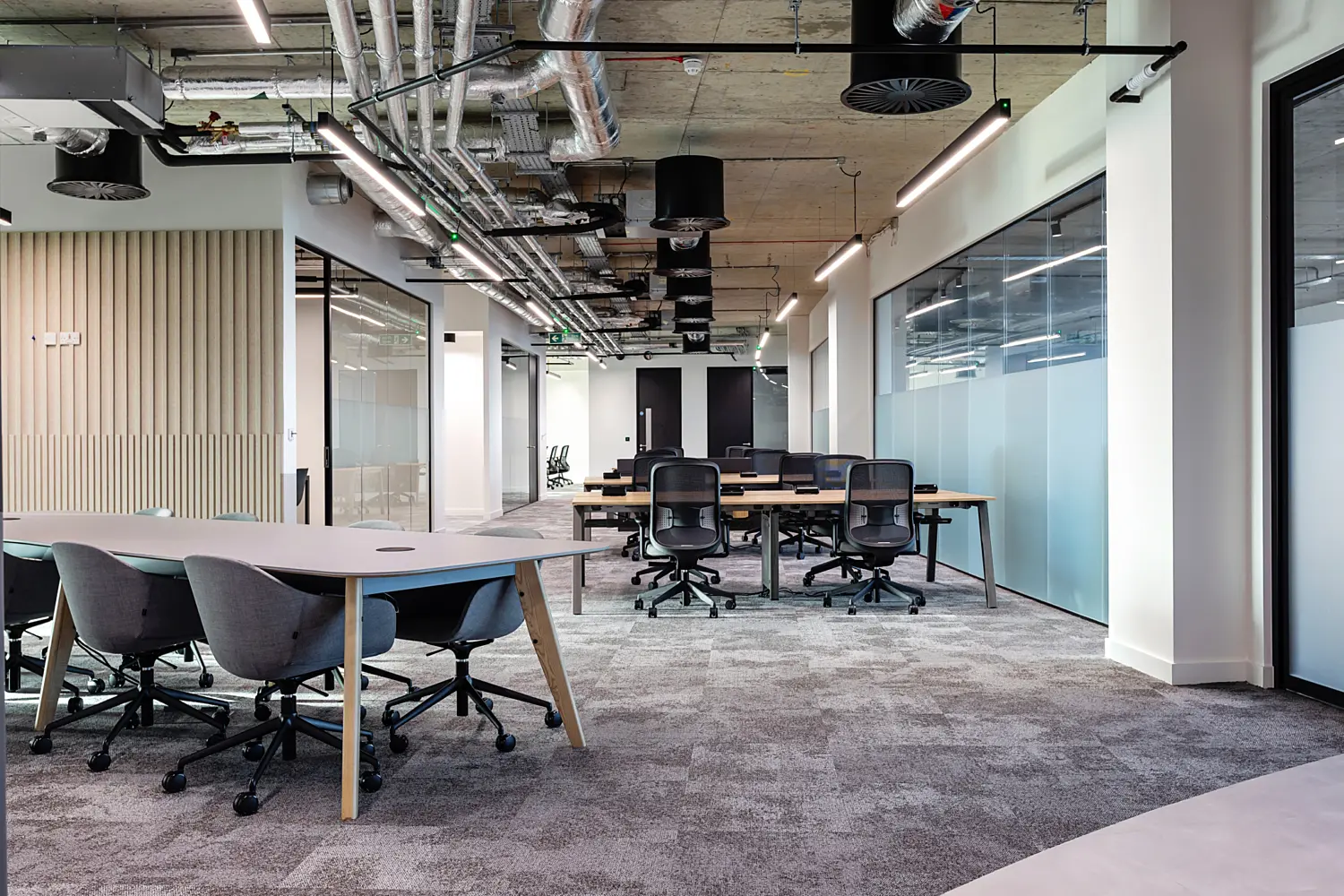 Open plan office workspace with a modern industrial style ceiling featuring exposed ductwork, pipes and linear lighting. In the centre and background, shared bench desks with light wood tops are arranged in rows, paired with black mesh office chairs on wheels. In the foreground, a rounded meeting table with a pale surface is surrounded by upholstered task chairs. Along the right side, a glass partition with a frosted lower section separates meeting rooms or private offices. On the left, a wall with vertical wooden slats adds texture near an open doorway. The floor is finished with patterned grey carpet tiles, and the space is lit with a mix of suspended strip lights and ceiling mounted fixtures.
