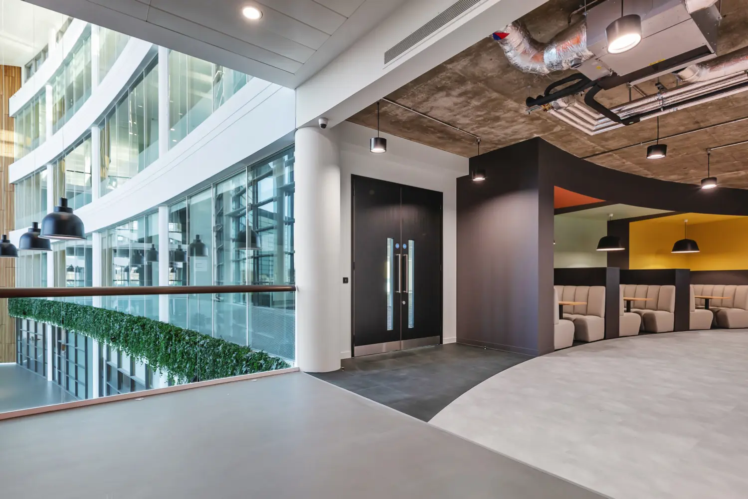 Office breakout and circulation area located beside a large curved glazed atrium wall that brings in natural light and views of adjacent floors. On the left, a glass balustrade with a timber handrail overlooks the atrium, with integrated greenery running along its base.  In the centre, a pair of black double doors sits within a dark wall section beneath an exposed industrial ceiling featuring visible ductwork, pipes and suspended lighting.  On the right, a recessed seating zone contains a row of upholstered booth seating in neutral tones, each paired with small tables and set within warm coloured alcoves, including yellow and orange. Pendant lights hang above each booth.  The flooring transitions from a darker surface near the doors to a lighter, smooth finish in the main walkway, complementing the clean white walls and contrasting with the raw concrete ceiling above.