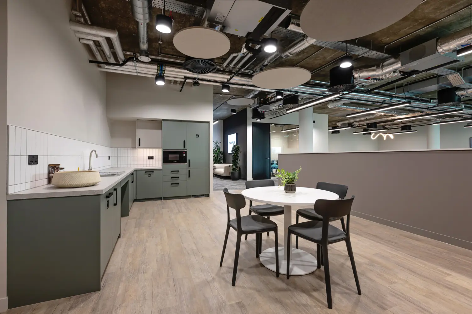 Modern office kitchenette and breakout area with an industrial style ceiling featuring exposed ductwork, pipework and circular acoustic panels suspended between lighting fixtures. In the foreground, a small round white table is paired with four dark chairs, with a potted plant placed at the centre.  To the left, a compact kitchenette runs along the wall with muted green cabinetry, a white tiled splashback, integrated appliances and a sink with a light coloured worktop. The flooring is finished in light wood effect planks, adding warmth to the space.  In the background, a half height partition separates the kitchenette from the wider office area, where additional seating and a sculptural light feature are visible. Neutral walls and clean lines contrast with the exposed industrial ceiling above, creating a balanced and contemporary look.