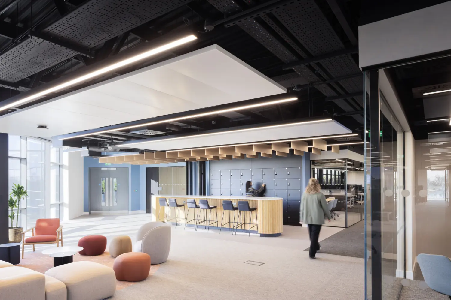 A bright open office space with soft seating in various colours arranged near full height windows. A bar height counter with stools sits in the centre of the room with a wall of lockers behind it. One person stands at the counter and another walks across the space. The ceiling features exposed black panels and linear lighting. A glass meeting room is visible on the right.
