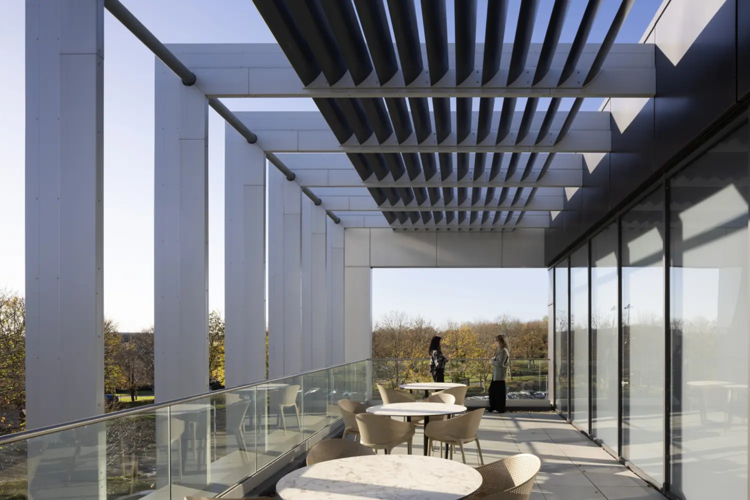 A terrace with round café tables and light chairs arranged along a glass balustrade. The space is partially shaded by a series of horizontal louvres overhead, which cast linear shadows across the floor. Floor to ceiling glazing runs along the right side of the terrace, reflecting the outdoor space. Beyond the terrace, trees and open landscape are visible. Two people stand near the far end of the terrace.