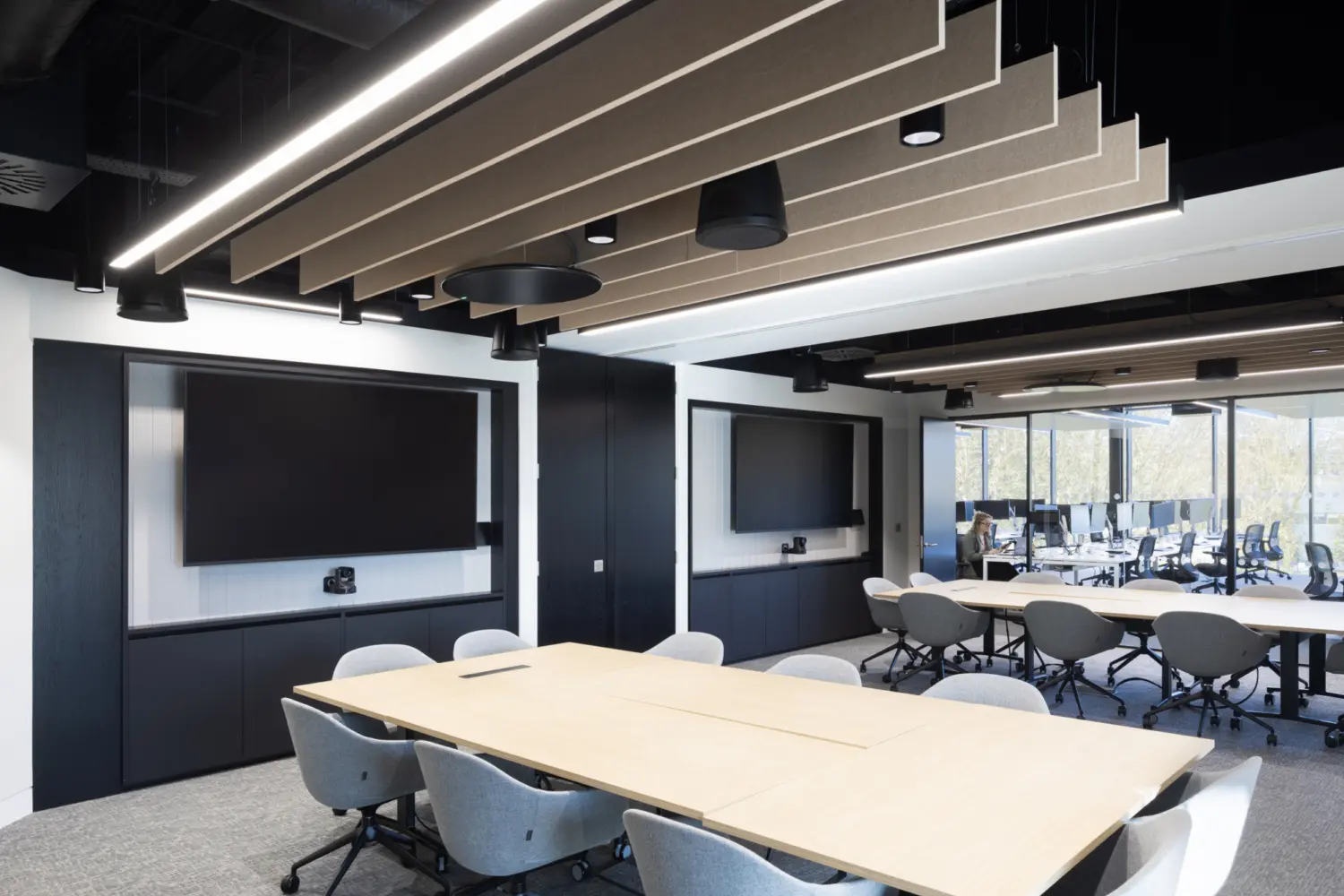 A modern meeting room with a large rectangular table surrounded by upholstered swivel chairs. Two large wall‑mounted screens sit on opposite walls above built‑in black cabinetry. Suspended acoustic panels and linear lighting are arranged across the ceiling. A second table with matching chairs is visible in the background near full height windows that look out onto trees.