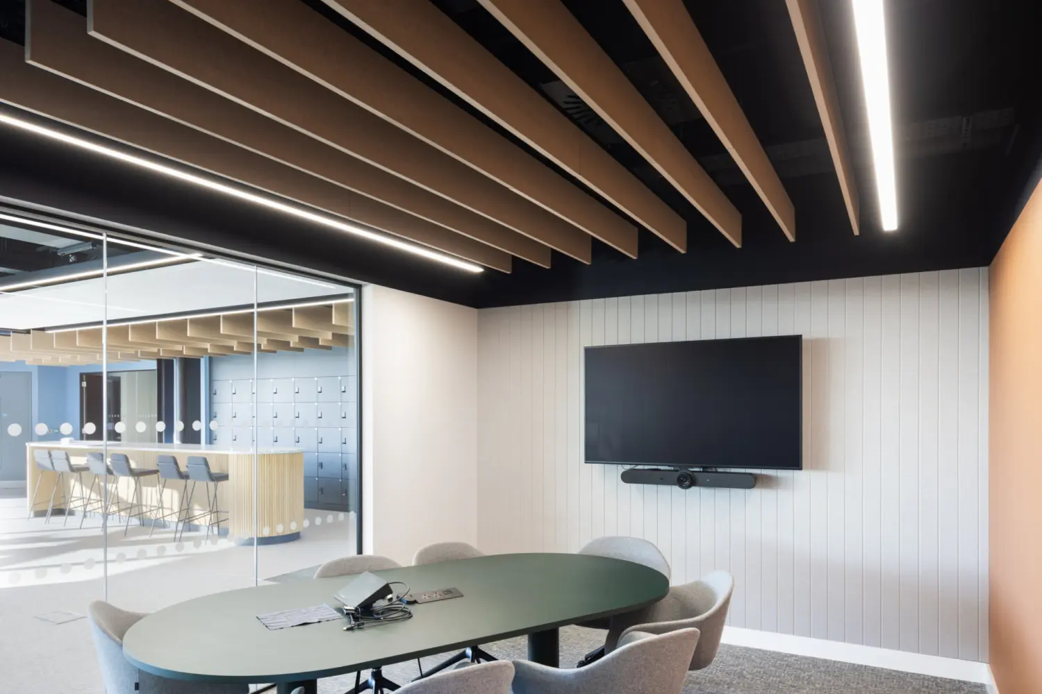 A small meeting room with an oval table surrounded by six upholstered swivel chairs. A large wall‑mounted screen and soundbar sit on a ribbed white feature wall, with an orange accent wall to the right. Suspended acoustic baffles run across the ceiling with linear lights fitted between them. A full height glass wall on the left looks out into an open workspace with a counter and stools visible in the background.
