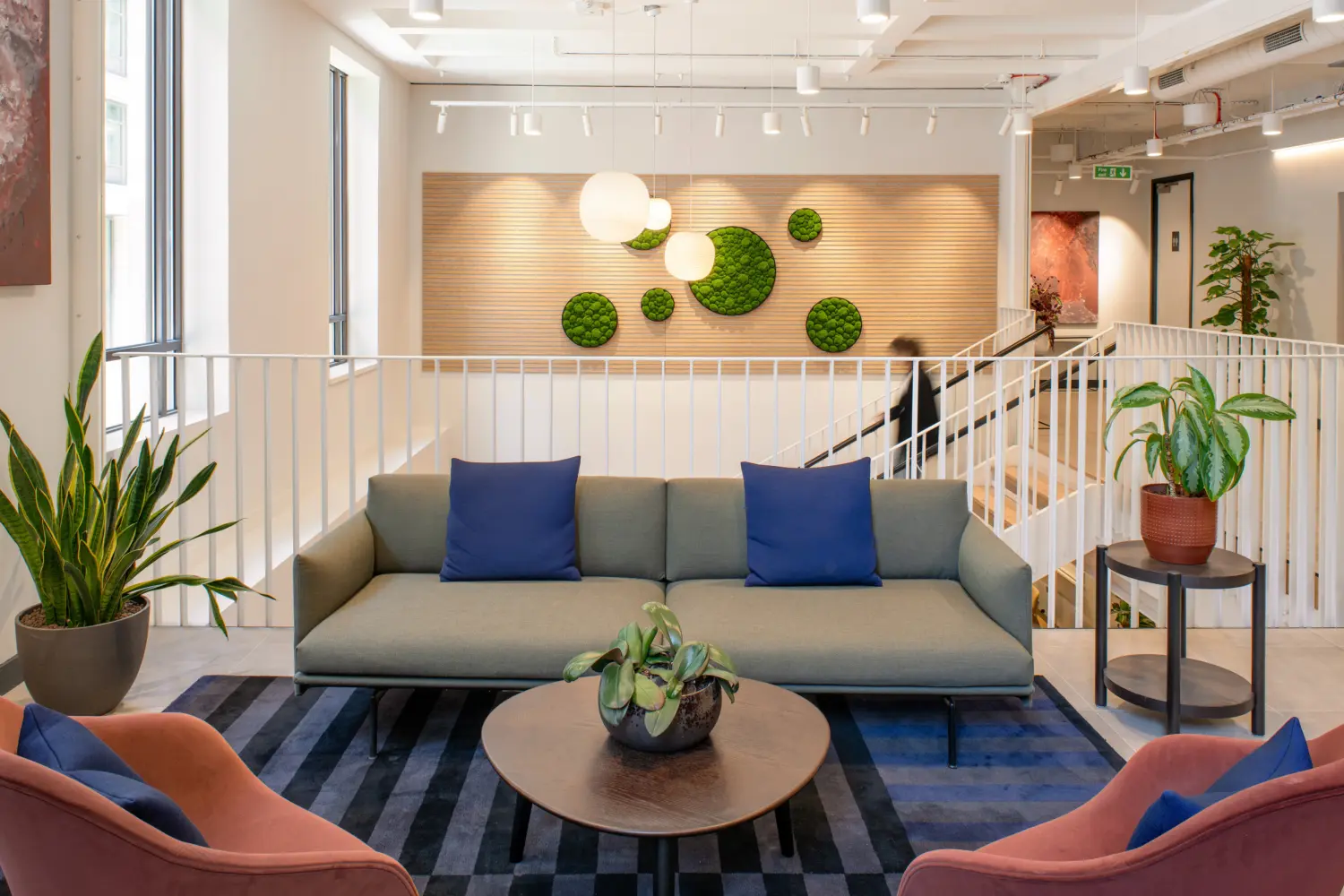 Contemporary office lounge area featuring a grey sofa with blue cushions positioned around a round coffee table on a patterned rug. Two upholstered lounge chairs and several indoor plants frame the seating area. In the background, a timber slat feature wall is decorated with circular green moss panels and illuminated by pendant globe lights. White railings overlook a staircase, while exposed ceilings, track lighting and large windows contribute to a bright, modern workplace environment. A blurred person is visible near the staircase in the background.