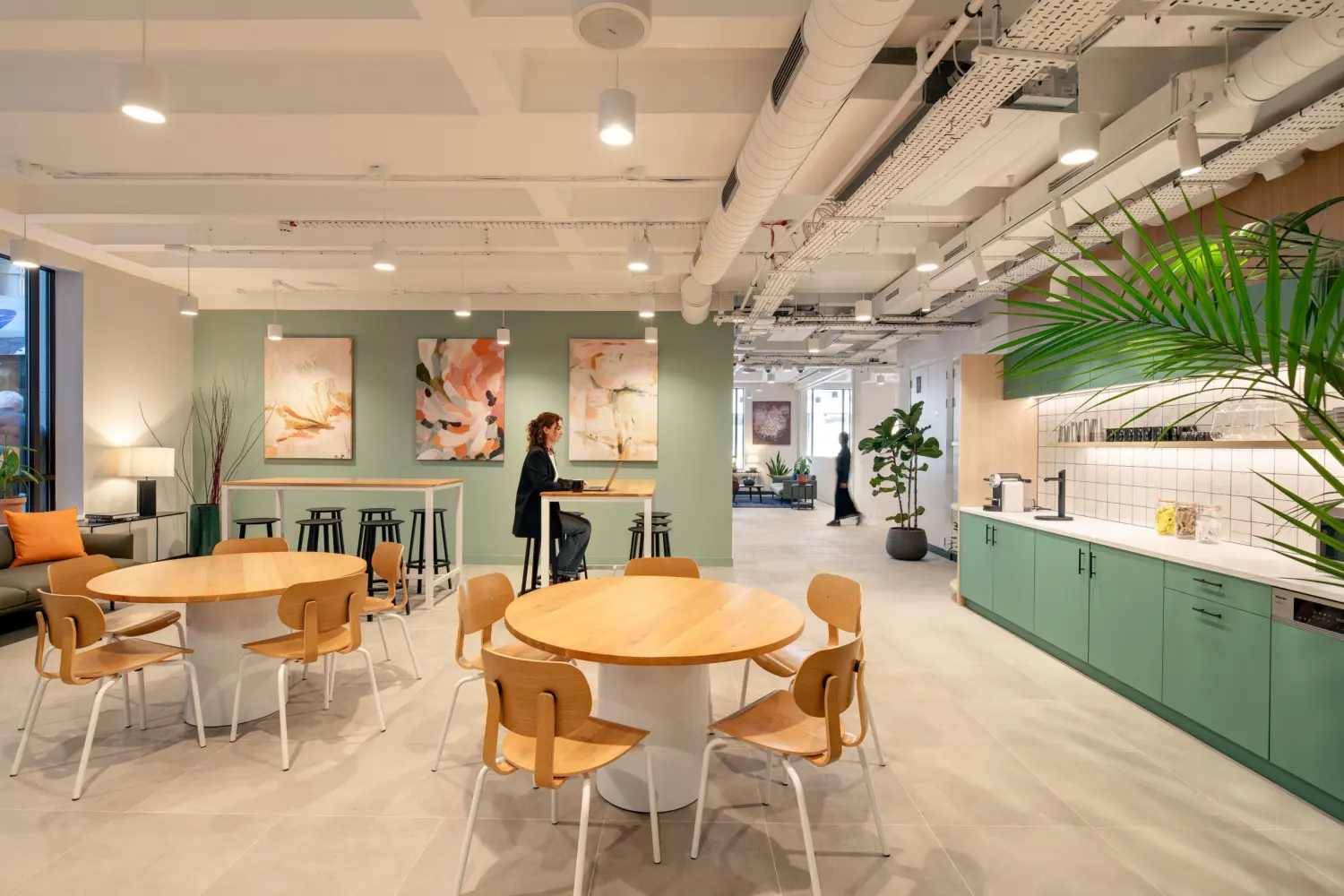Contemporary office breakout area with round wooden tables and matching chairs arranged throughout an open-plan space. A kitchenette with sage green cabinetry, white worktops and a tiled splashback runs along the right-hand side, while high benches and stools provide additional seating against a green feature wall decorated with abstract artwork. Exposed ceilings, suspended lighting and indoor plants contribute to a modern workplace aesthetic. Two people are visible in the space, one seated at a high table and another walking in the background.