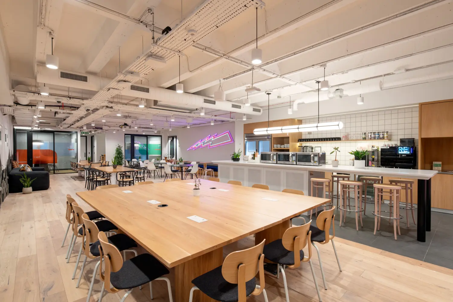 Open-plan office breakout and dining area featuring a large communal wooden table with integrated power points surrounded by chairs. A spacious kitchenette with a long white breakfast bar, stools, coffee machines, microwaves and tiled splashback runs along the right-hand side. Additional café-style tables and lounge seating are arranged throughout the space, while a purple neon wall feature adds a vibrant focal point in the background. Exposed ceilings, suspended lighting, timber finishes and indoor plants create a bright, contemporary workplace environment.