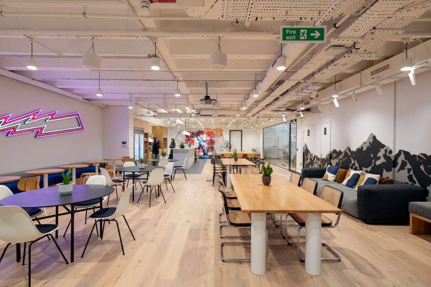 Large open-plan office breakout space with a mix of dining, collaboration and lounge areas. Round café-style tables and chairs are arranged on the left, while a long communal wooden table with woven-back chairs runs through the centre. A lounge seating area lines the right-hand wall beneath a black mountain-themed mural, and a neon wall feature provides a colourful focal point on the left. The space extends towards a kitchenette and a vibrant graffiti-style mural in the distance. Exposed ceilings, suspended lighting, glazed meeting rooms and indoor plants contribute to a bright, contemporary workplace environment.