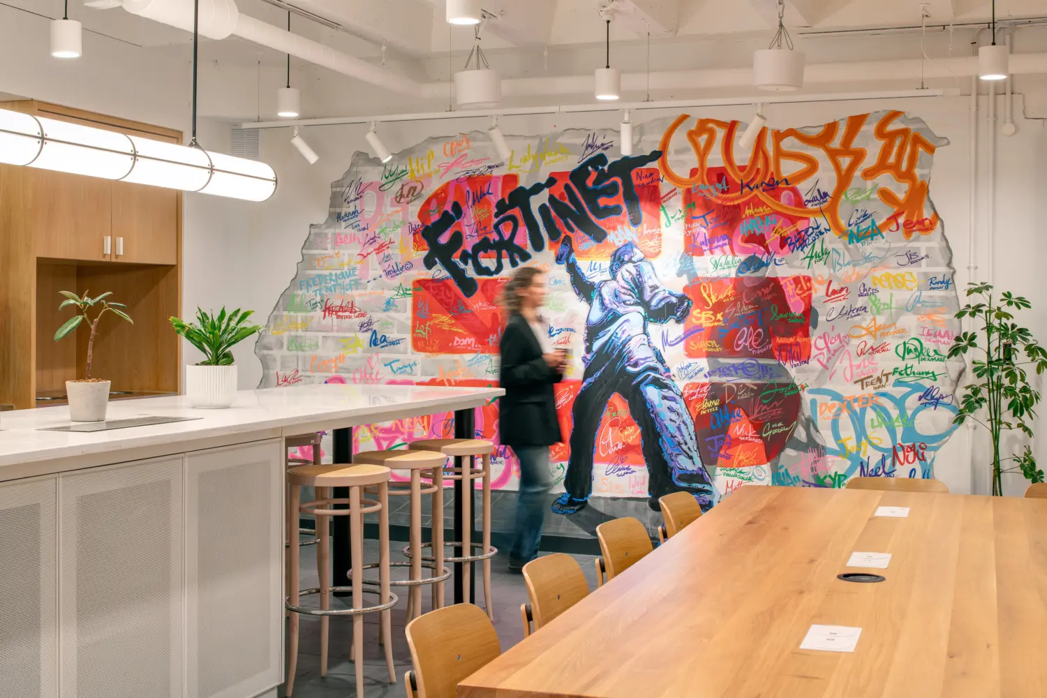 Modern breakout and collaboration space featuring a large colourful graffiti-style mural with the word "Fortinet" and numerous handwritten signatures across a faux exposed-brick wall. A long timber meeting table with integrated power points occupies the foreground, while a high counter with stools sits to the left. Indoor plants, timber cabinetry and suspended pendant lighting contribute to a bright contemporary office environment. A person walking through the space appears blurred due to movement.