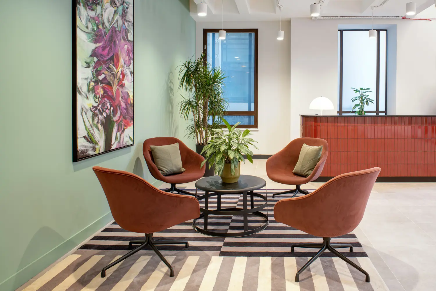 Contemporary office lounge area featuring four upholstered terracotta-coloured lounge chairs arranged around a round black coffee table with a plant at the centre. The seating area sits on a striped rug and is framed by indoor plants, a sage green feature wall and a large floral artwork. In the background, a reception desk with a red ribbed front panel is positioned beneath pendant lighting. Large windows and a light, neutral colour palette create a bright and welcoming workplace environment.