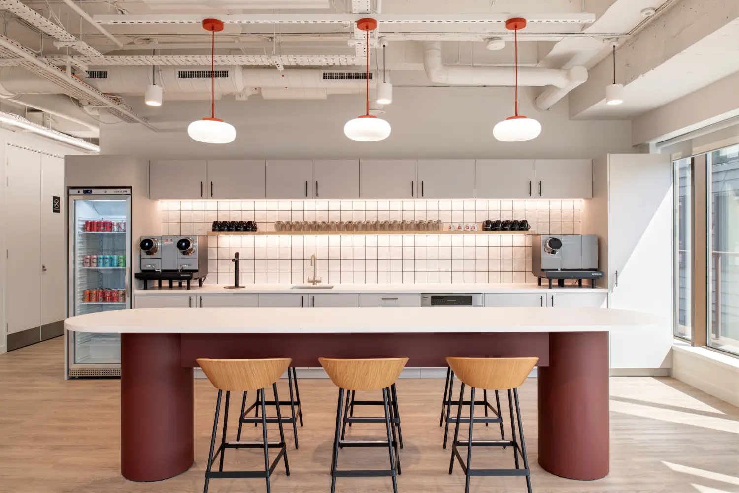 Contemporary office kitchenette featuring a large white island with three wooden bar stools and a deep red base. White cabinetry lines the back wall above a tiled splashback, with coffee machines, glassware and a sink arranged along the countertop. A drinks fridge stocked with canned beverages stands to the left, while floor-to-ceiling windows on the right bring natural light into the space. Exposed services, white-painted ceilings and pendant lights with red accents create a modern industrial-style aesthetic.