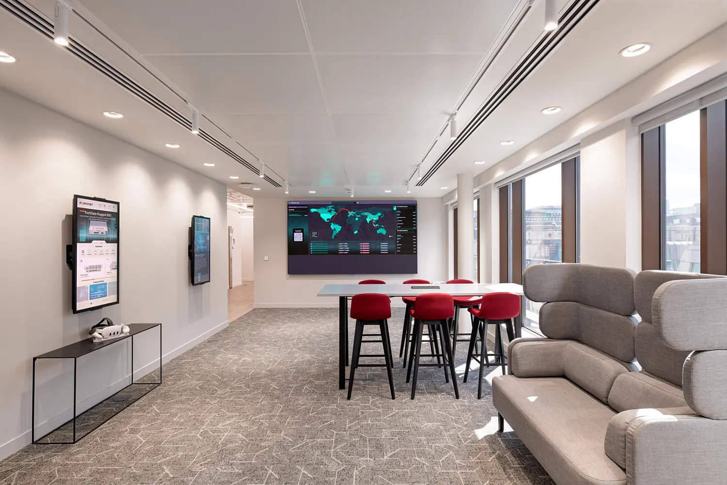 Modern office collaboration space featuring a high communal table with red upholstered stools positioned in front of a large digital display showing a world map and cybersecurity monitoring dashboard. Floor-to-ceiling windows along the right-hand side provide natural light and city views, while a high-backed grey lounge sofa offers informal seating in the foreground. Framed technology displays are mounted on the left wall, and recessed lighting and a neutral colour palette create a bright, professional workplace environment.