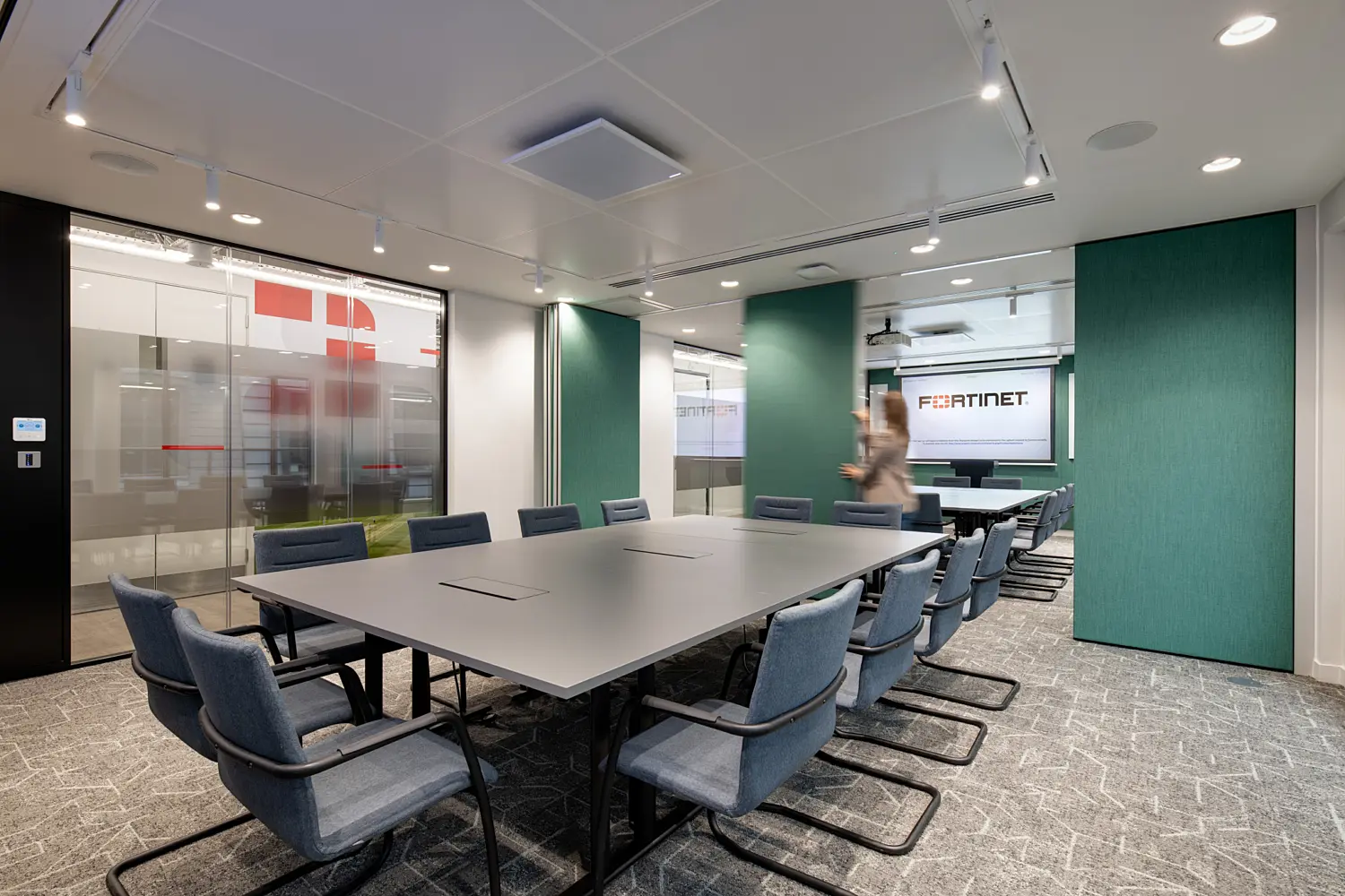 Modern boardroom featuring a large rectangular conference table surrounded by blue upholstered meeting chairs. Glazed partitions with Fortinet branding line the left-hand side, while green acoustic wall panels add colour and texture to the space. At the far end of the room, a presentation screen displays the Fortinet logo. Recessed and track lighting provide even illumination, and a blurred person walking near the screen adds a sense of activity within the contemporary office environment.