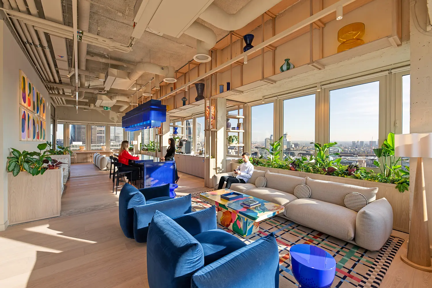 Modern office lounge with floor-to-ceiling windows overlooking the City of London skyline. The space features a large curved beige sofa, two blue lounge chairs, a colourful coffee table and abundant indoor plants. A glossy blue bar and matching suspended light fixture sit at the centre, while decorative glass vases line open shelving above. Three people occupy the space, with two seated at the bar and one seated on the sofa. Exposed ceilings, light wood finishes and natural daylight create a bright, contemporary workplace setting.