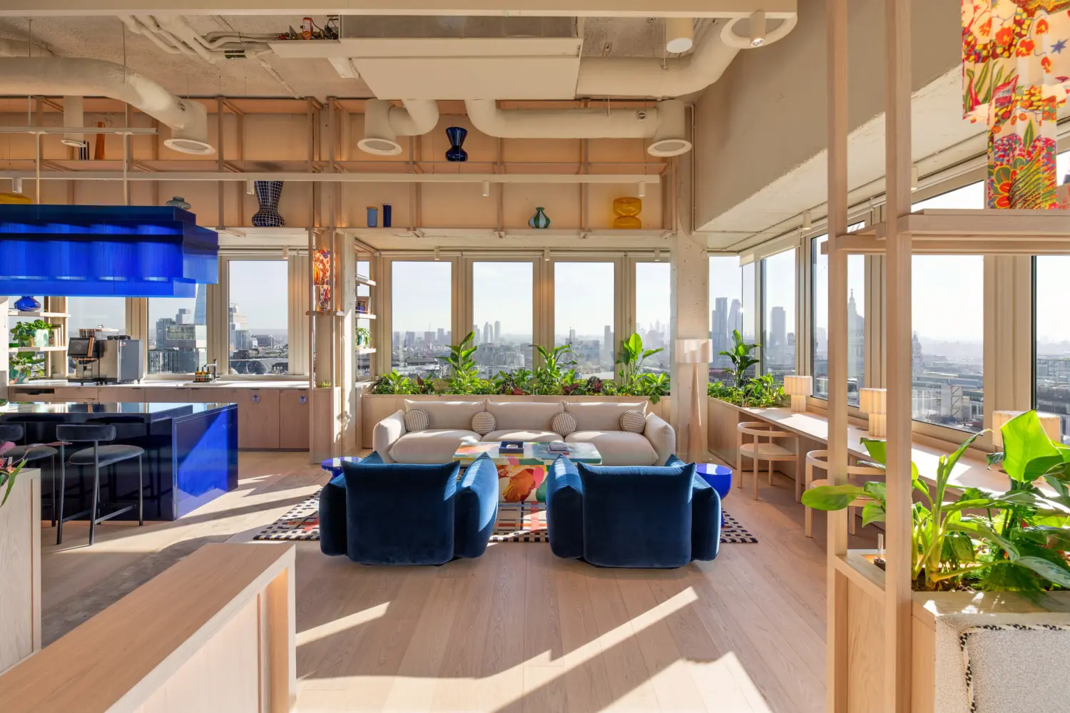 Contemporary office lounge with panoramic views across the London skyline through floor-to-ceiling windows. The space features a seating area with a curved beige sofa, four blue lounge chairs and a colourful coffee table set on a patterned rug. A glossy blue bar with matching pendant lighting sits to the left, while open timber shelving displays decorative glassware and integrated planters filled with greenery. Natural daylight floods the interior, complementing the light wood finishes, exposed ceiling services and vibrant design details throughout the workplace.