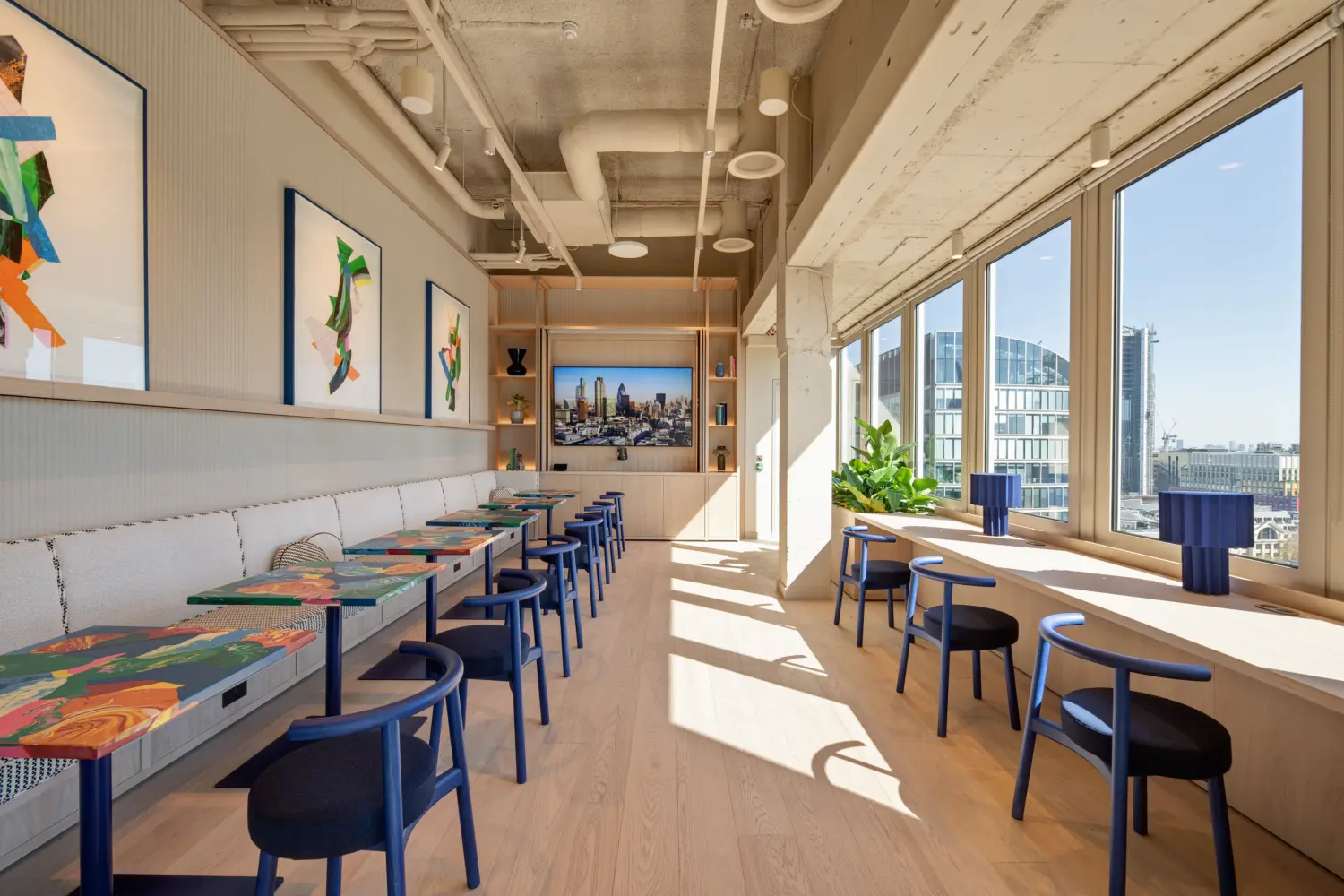 Contemporary office touchdown spaces with floor-to-ceiling windows offering panoramic city views. A row of colourful tables with blue chairs runs alongside upholstered banquette seating, while a window-facing counter provides additional seating for informal work or meetings. Framed abstract artwork decorates the wall, and a large display screen at the far end shows a London skyline image. Abundant natural daylight fills the space, complementing the light wood finishes, exposed ceiling services and modern workplace design.
