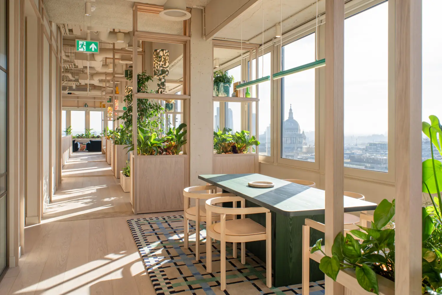 Sunlit office corridor with a meeting booth positioned beside floor-to-ceiling windows overlooking the London skyline, including a distant view of St Paul's Cathedral. The booth features a green table, light wood chairs and a patterned rug, framed by open timber shelving filled with plants. Planters line the walkway, creating a biophilic environment throughout the space. Natural light casts long shadows across the pale timber flooring, while exposed ceilings and light wood finishes contribute to the contemporary workplace design.