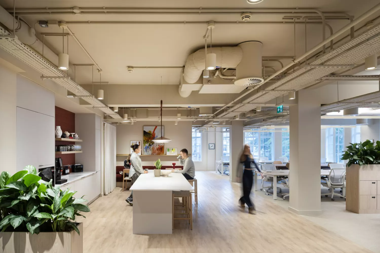 Modern open plan office with a bright, minimalist design featuring a central communal table with high stools, integrated coffee station and natural wood flooring. Exposed ceiling services and suspended lighting create an industrial aesthetic, complemented by large windows for natural light and greenery for a biophilic touch. Flexible workstations are visible in the background, promoting collaboration and contemporary workplace design.