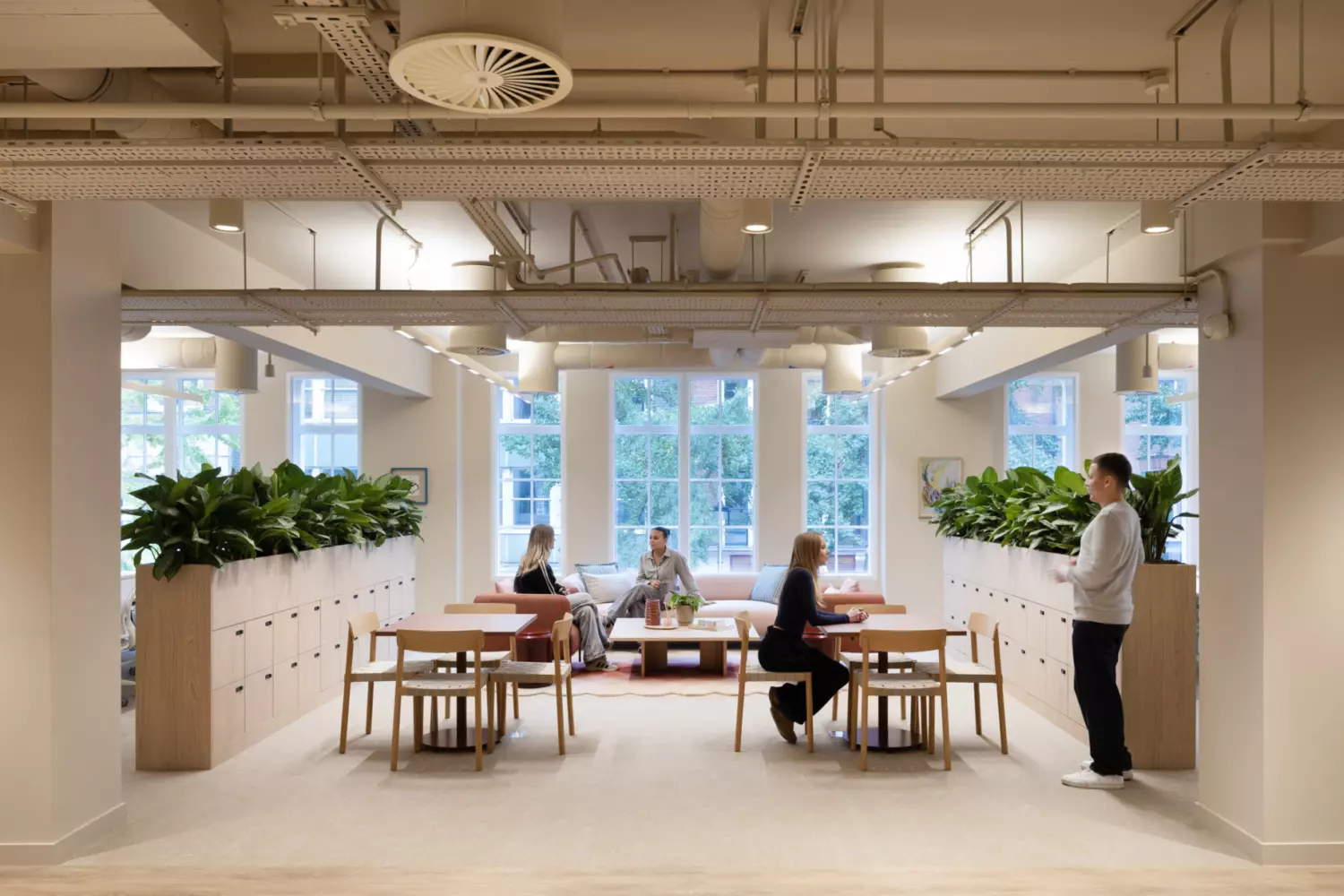 Collaborative office lounge with natural light from large windows, featuring two wooden tables with chairs and a central soft seating area. Green plants line storage units on both sides, adding a biophilic element. Exposed ceiling services and neutral tones create a modern, open-plan design that encourages informal meetings and flexible working