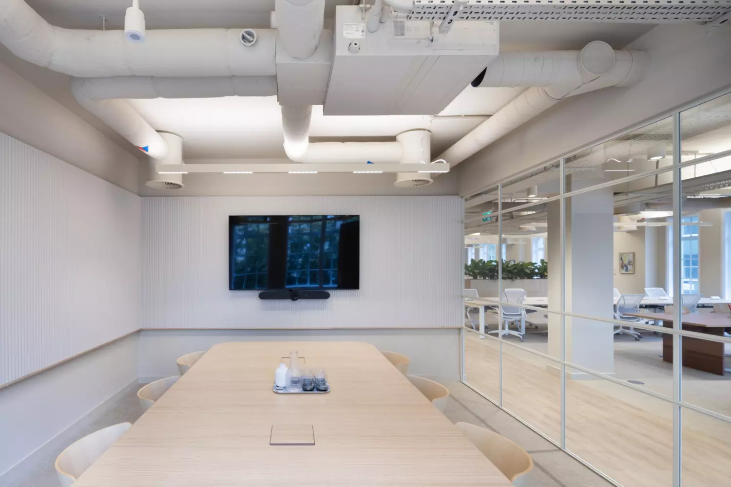 Contemporary meeting room with a large wooden conference table and six chairs, featuring a wall-mounted screen for presentations. The space has exposed ceiling ducts and cable trays for an industrial look, neutral wall panels for acoustic control, and glass partitions that open into an adjacent open-plan office with natural light and greenery