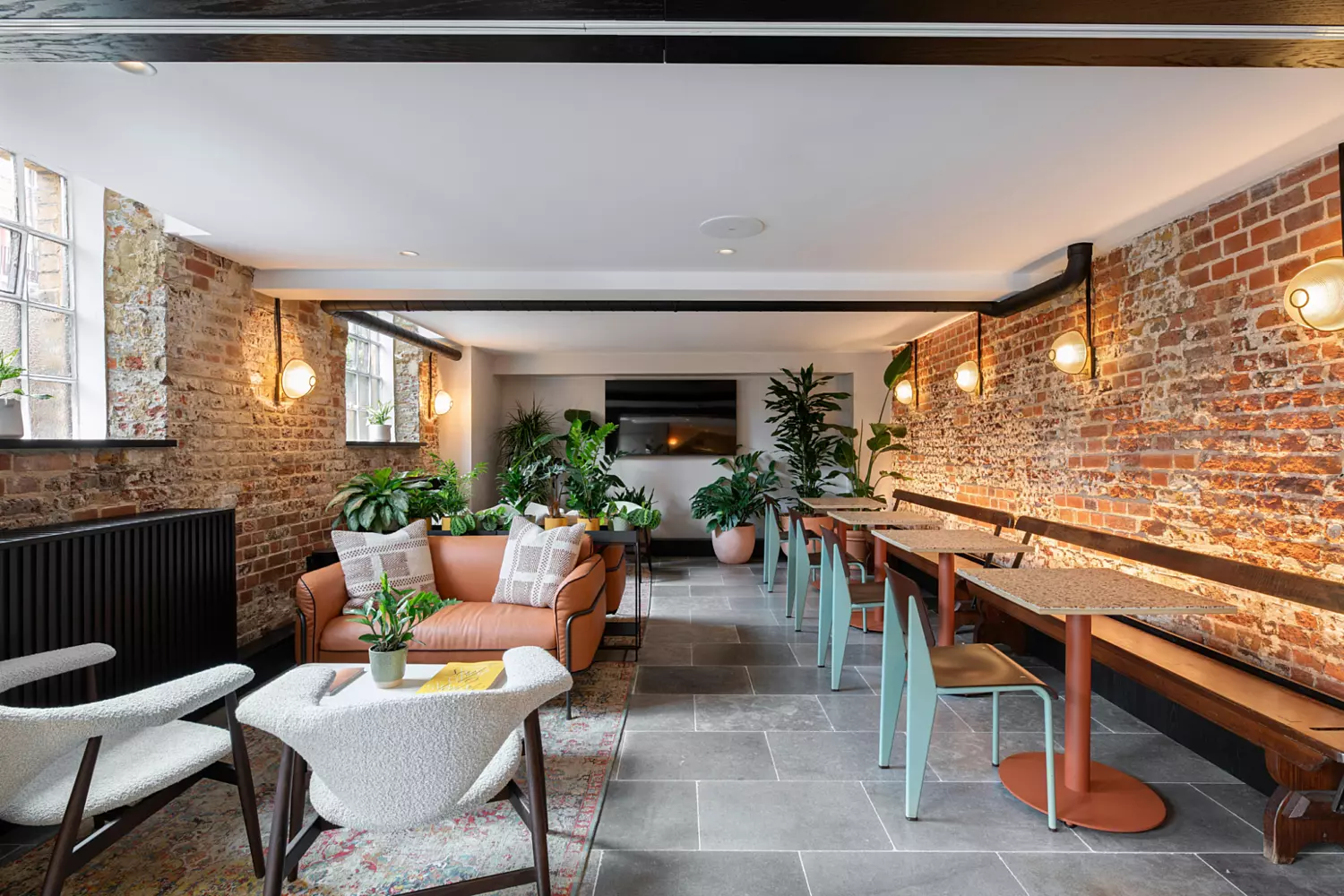 A well-lit room with exposed brick walls and large windows, combining contemporary and vintage furnishings. The space includes a brown leather sofa, white armchairs, and wooden tables paired with metal chairs. Large grey floor tiles cover the room, while potted plants add touches of greenery. Hanging light fixtures are mounted along the brick wall, contributing to the room’s warm and eclectic character.