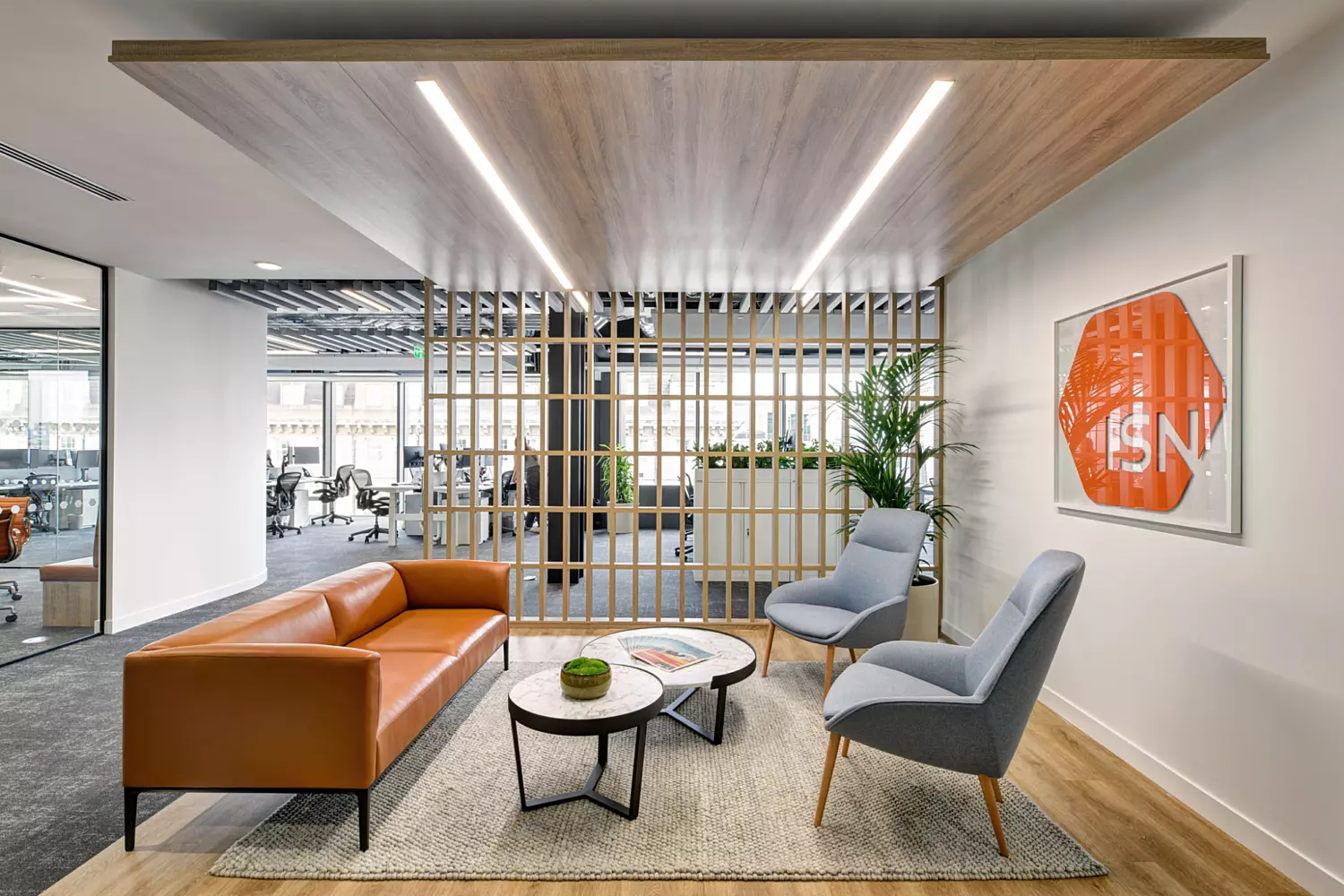 An office lounge area featuring a brown leather sofa, two grey armchairs, and a pair of round coffee tables placed on a beige rug. A wooden slatted partition provides a visual divide from the open-plan office space beyond, which includes desks and chairs. The ceiling is fitted with recessed lighting, and a framed artwork with an orange geometric design and the text "ISN" hangs on the wall, adding a bold visual accent.