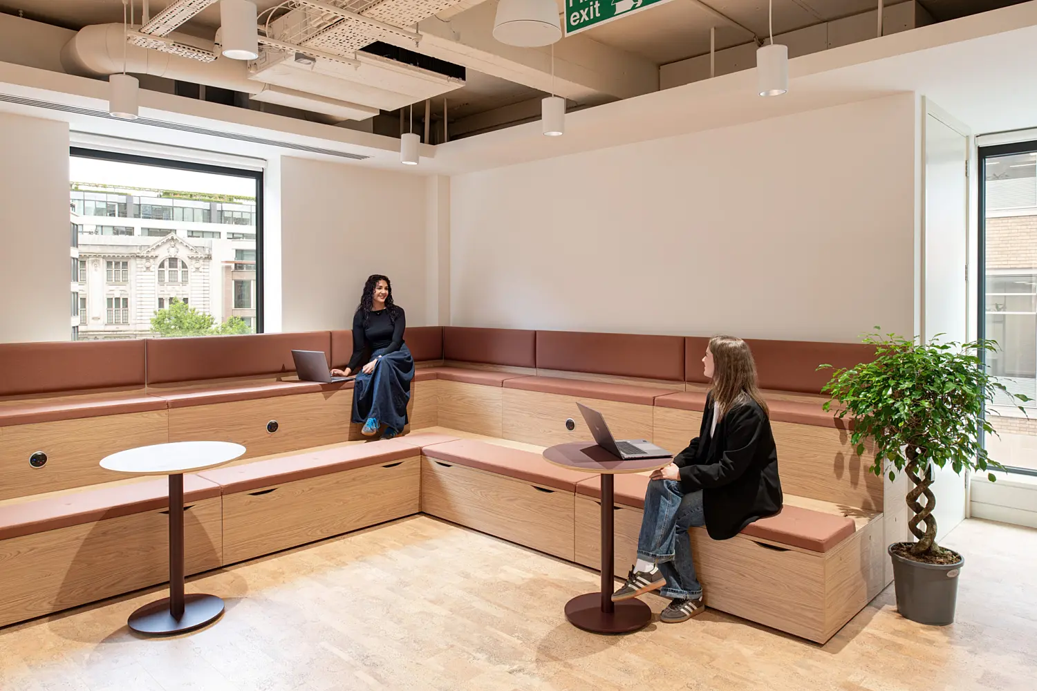 Tiered breakout seating area with integrated timber benches, upholstered cushions and small round tables designed for informal meetings and focused work. Two people use laptops in the space, which is illuminated by natural light from large windows overlooking surrounding office buildings. Warm timber finishes, contemporary lighting and indoor planting create a bright, welcoming workplace environment.