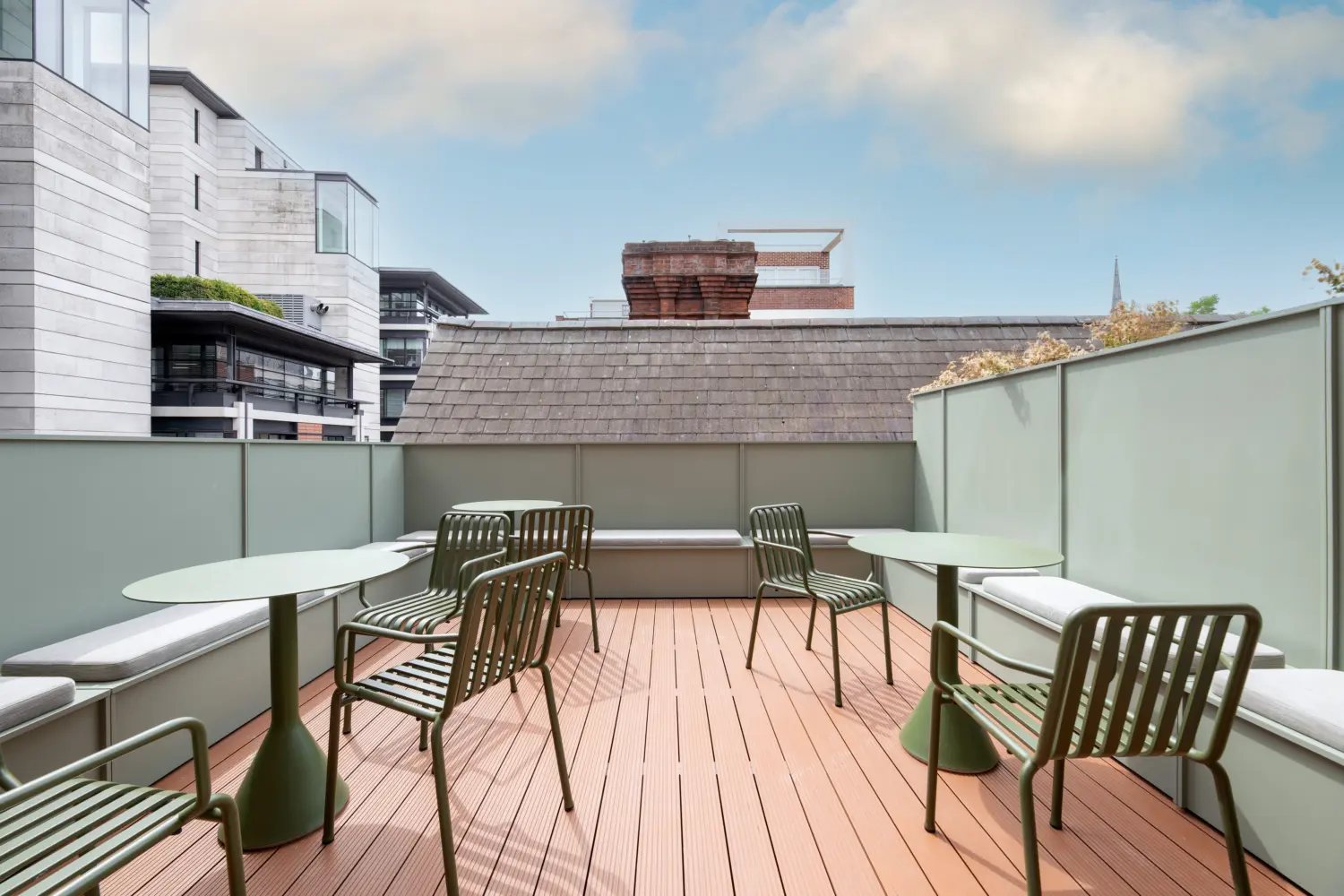 Outdoor roof terrace featuring built-in bench seating, round tables and metal chairs arranged for informal meetings and relaxation. Timber-effect decking and pale green privacy screens create a calm, enclosed setting, while surrounding office buildings and a blue sky provide an urban backdrop. The space offers a contemporary outdoor area for dining, socialising and collaborative work.