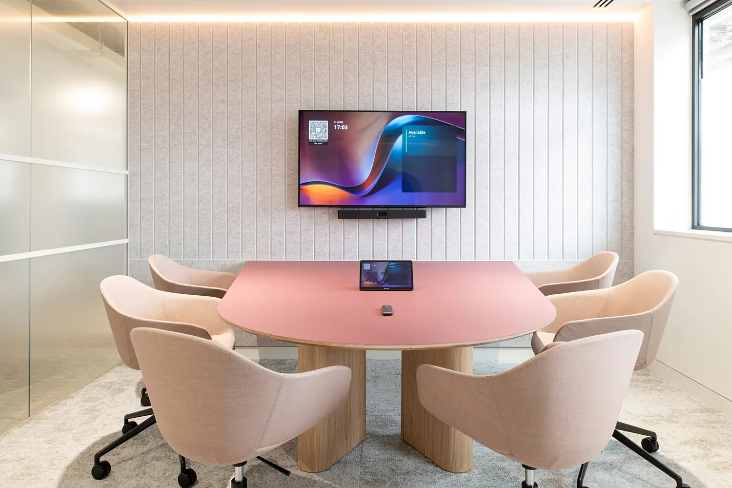 Small meeting room featuring a curved blush-pink conference table surrounded by six upholstered chairs on castors. A wall-mounted display screen and video conferencing equipment provide facilities for hybrid meetings, while a textured acoustic feature wall with integrated lighting creates a refined backdrop. Glazed partitions and large windows bring natural light into the space, contributing to a bright, contemporary workplace setting.