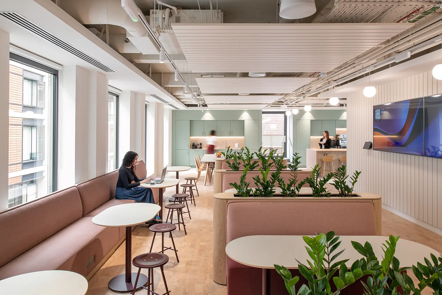 Contemporary office café and breakout area with banquette seating, round tables and integrated planters filled with greenery. Large windows bring natural light into the space, while a kitchenette and refreshment counter sit at the far end. A wall-mounted display screen, soft colour palette and exposed ceiling services create a modern, welcoming environment designed for informal meetings, dining and collaborative work.
