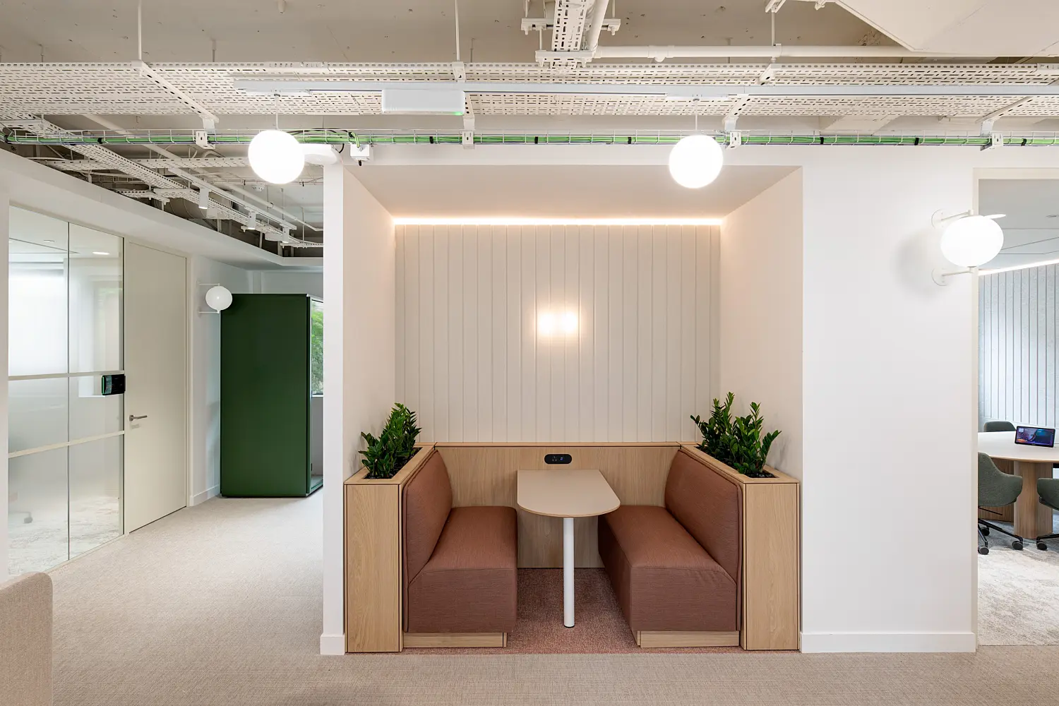 Semi-enclosed office booth with upholstered bench seating arranged around a small meeting table for informal discussions and focused work. Timber planters with integrated greenery frame the seating area, while a vertical acoustic feature wall with soft lighting creates a sense of privacy. Glazed meeting rooms and collaboration spaces surround the booth, and exposed ceiling services with globe pendant lights contribute to the contemporary workplace design.