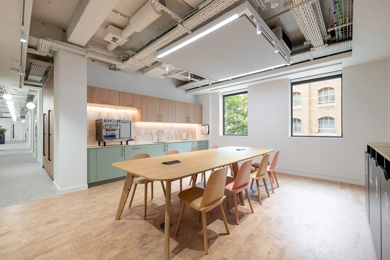 Office kitchen and dining area featuring a large communal table surrounded by mixed timber and pastel-coloured chairs. A kitchenette with integrated cabinetry, worktops and a coffee machine runs along one wall, while large windows bring natural light into the space and provide views of neighbouring buildings. Suspended acoustic panels, exposed ceiling services and warm timber finishes create a bright, contemporary environment for dining, informal meetings and collaboration.