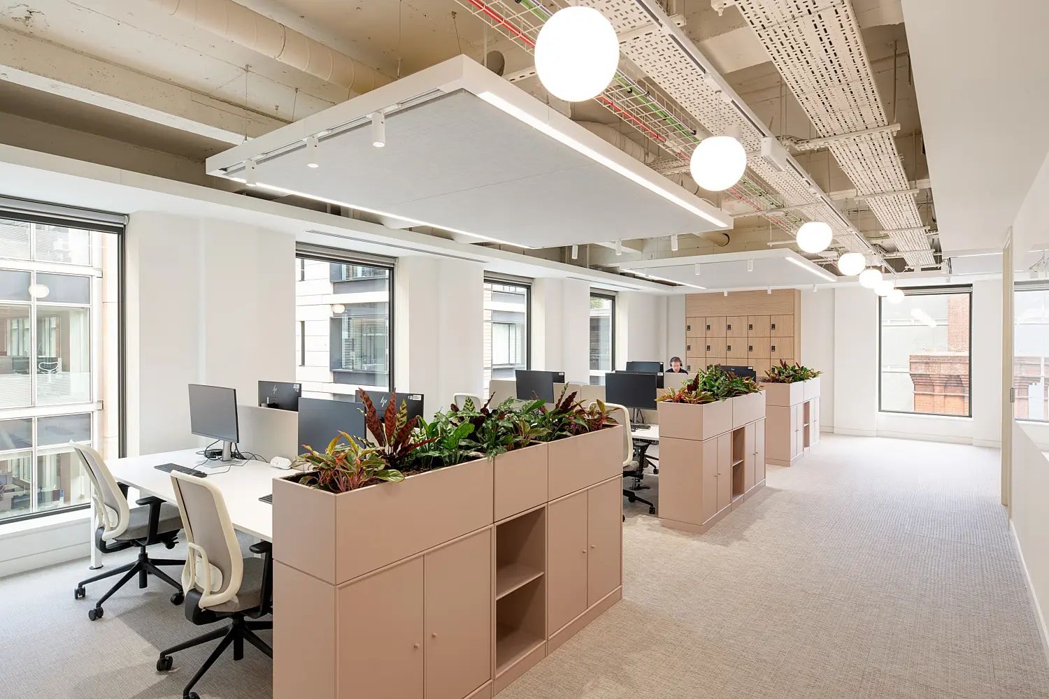Open-plan office workspace with bench-style desks, ergonomic task chairs and computer monitors arranged in clusters. Integrated planters filled with lush greenery divide the workstations, while large windows provide abundant natural light and views of neighbouring buildings. Suspended acoustic ceiling panels, globe pendant lights and exposed building services create a bright, contemporary workplace designed for focused work and collaboration.