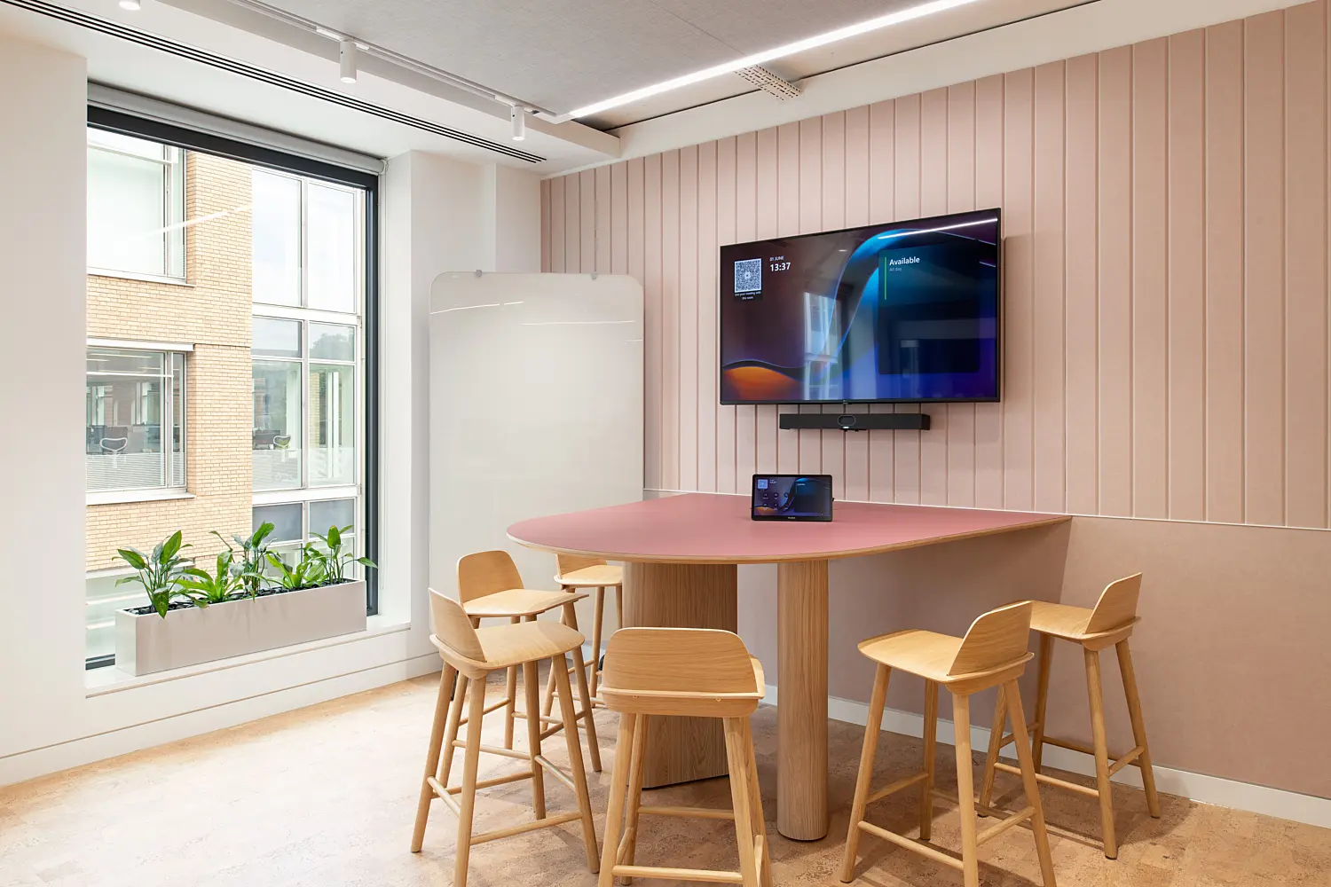 Small collaboration area featuring a high table with timber stools positioned beside a large window and indoor planter. A wall-mounted display screen and video conferencing equipment support informal meetings and hybrid working, while a soft pink acoustic feature wall provides a contemporary backdrop. The space is bright and welcoming, with natural light, warm timber finishes and clean, modern detailing throughout.