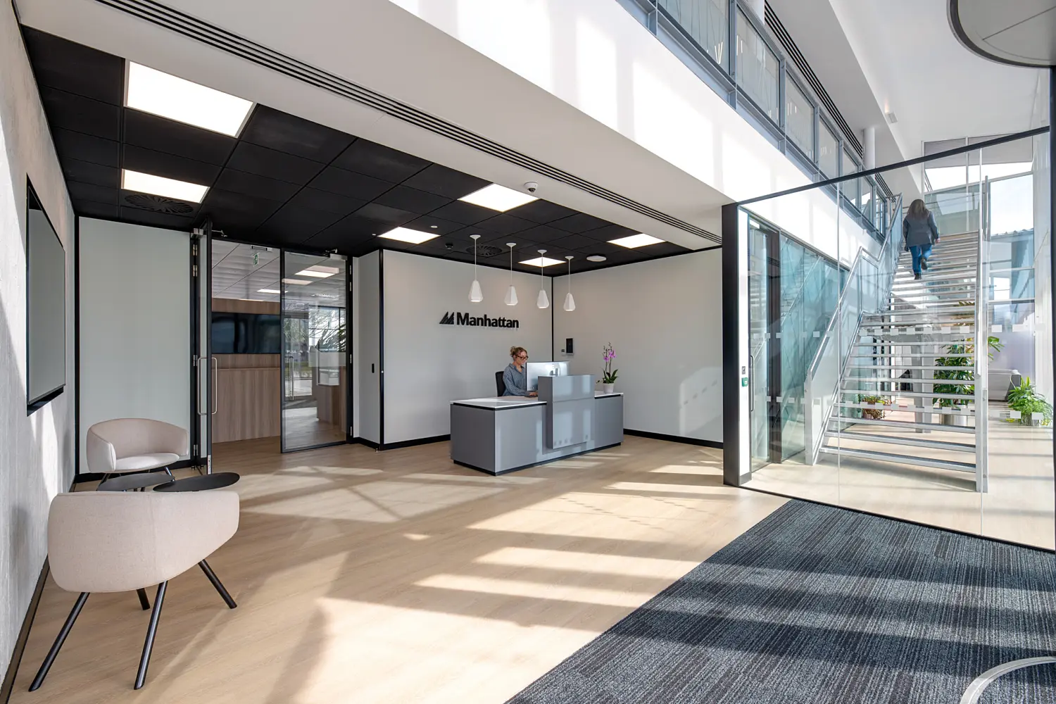 Modern office reception area with light wood flooring, a grey welcome desk and a feature wall displaying the Manhattan logo. A person is seated at the reception desk, while another person walks up a glass-sided staircase in a bright atrium. The space features full-height glazing, white walls, black ceiling panels with integrated lighting and contemporary lounge seating, with natural daylight creating strong shadows across the floor.