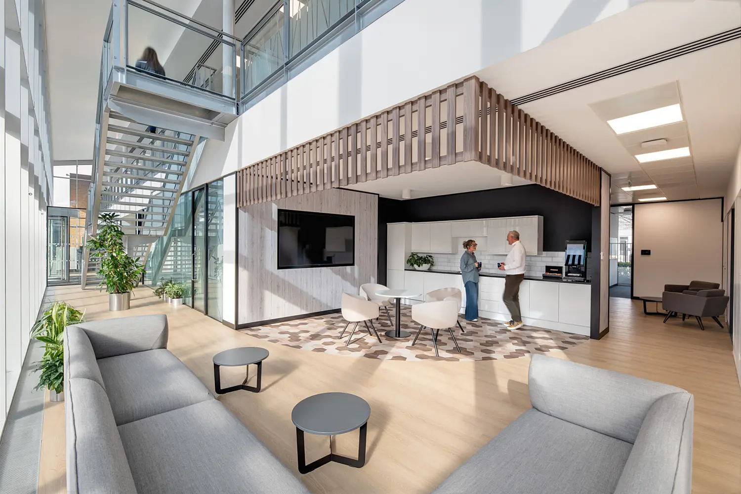 Bright, modern office breakout area with light wood flooring, grey sofas and small round tables. A kitchenette with white cabinetry sits beneath a timber slatted feature structure, while two people stand beside a coffee machine. A large wall-mounted screen, indoor plants and a glass-and-metal staircase leading to an upper level contribute to the open, airy design. Natural light streams through full-height windows along the left side of the space.