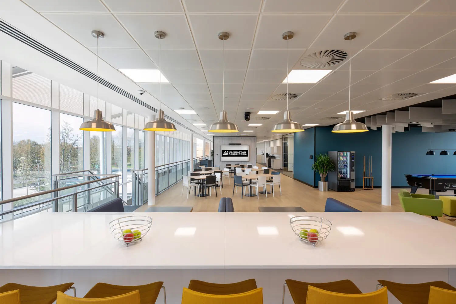 Spacious office breakout and social area featuring a long white breakfast bar with mustard-coloured stools, pendant lighting and light wood flooring. Floor-to-ceiling windows run along the left side, filling the space with natural daylight. In the centre, café-style tables and chairs face a feature wall displaying the Manhattan Associates logo. A vending machine, pool table and colourful lounge seating sit to the right, creating a multi-purpose area for dining, collaboration and relaxation.