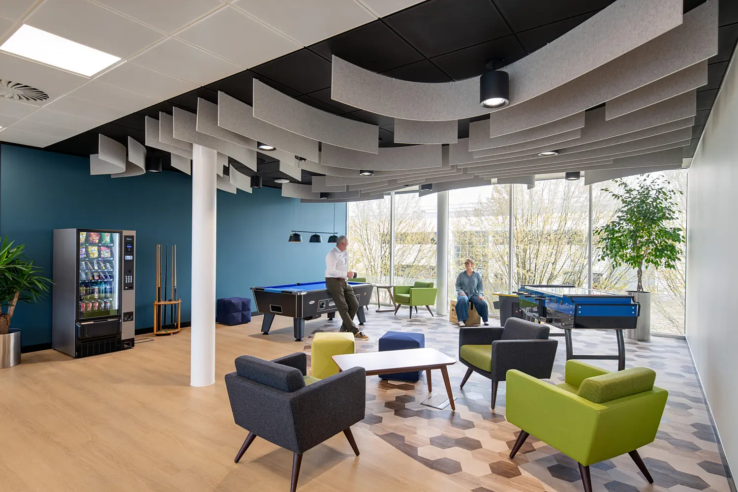 Office breakout and games area with a mix of lounge seating, pool tables and casual meeting spaces. Grey and green armchairs surround low tables on a patterned carpet, while two people are positioned near the games area. Floor-to-ceiling windows along the far wall provide natural daylight and views of trees outside. The space includes a vending machine, cue rack and feature acoustic ceiling baffles in a curved arrangement above, creating a bright and contemporary environment for relaxation and informal collaboration.
