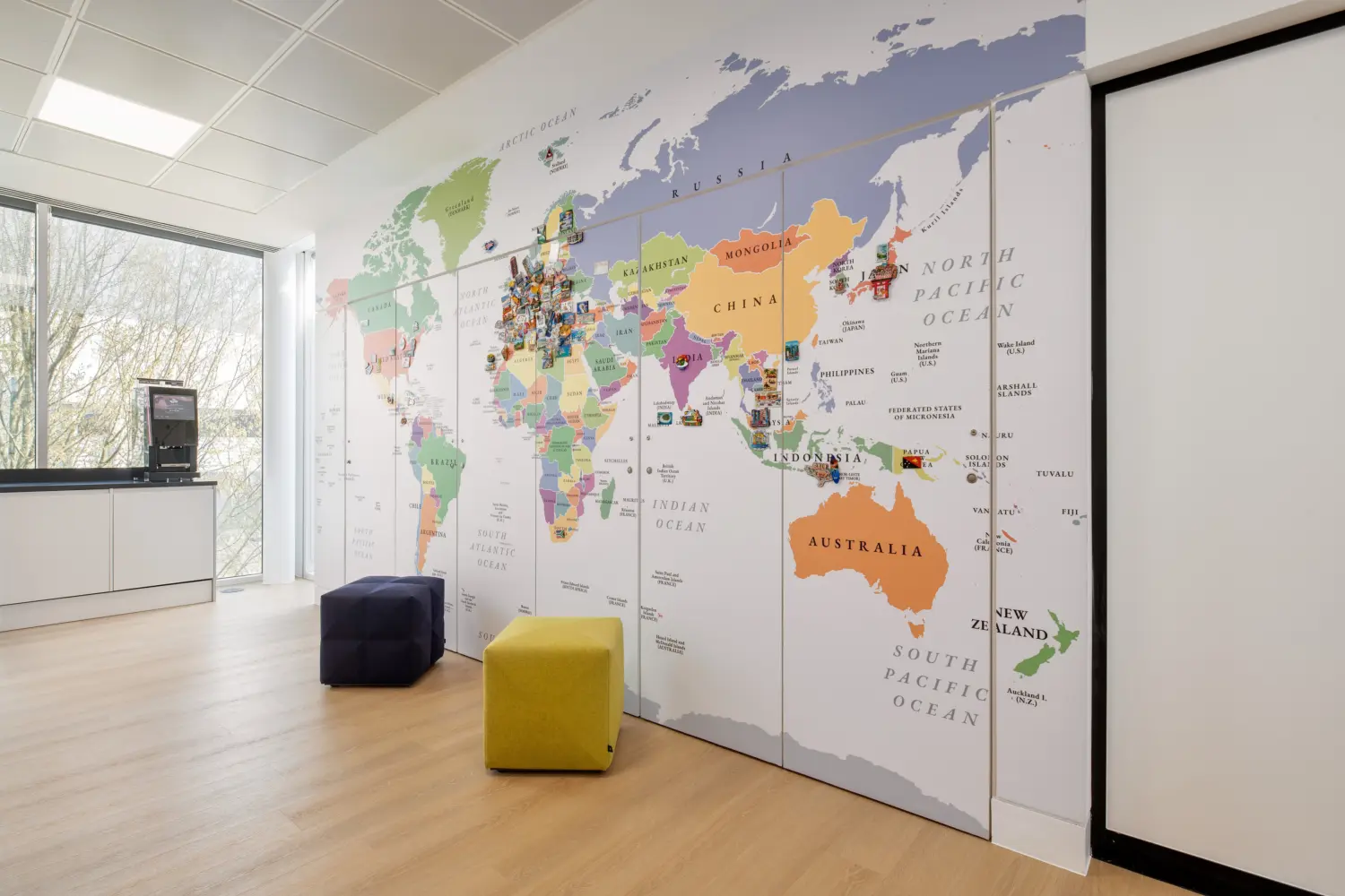 Bright office breakout area featuring a large floor-to-ceiling world map graphic displayed across a wall. Colourful markers and photographs are positioned on various countries, creating an interactive display. Two upholstered cube stools in yellow and dark blue sit on the light wood floor in front of the map. To the left, a refreshment station is positioned beside full-height windows that provide natural daylight and views of trees outside. The space combines collaborative features with clean, contemporary office design.