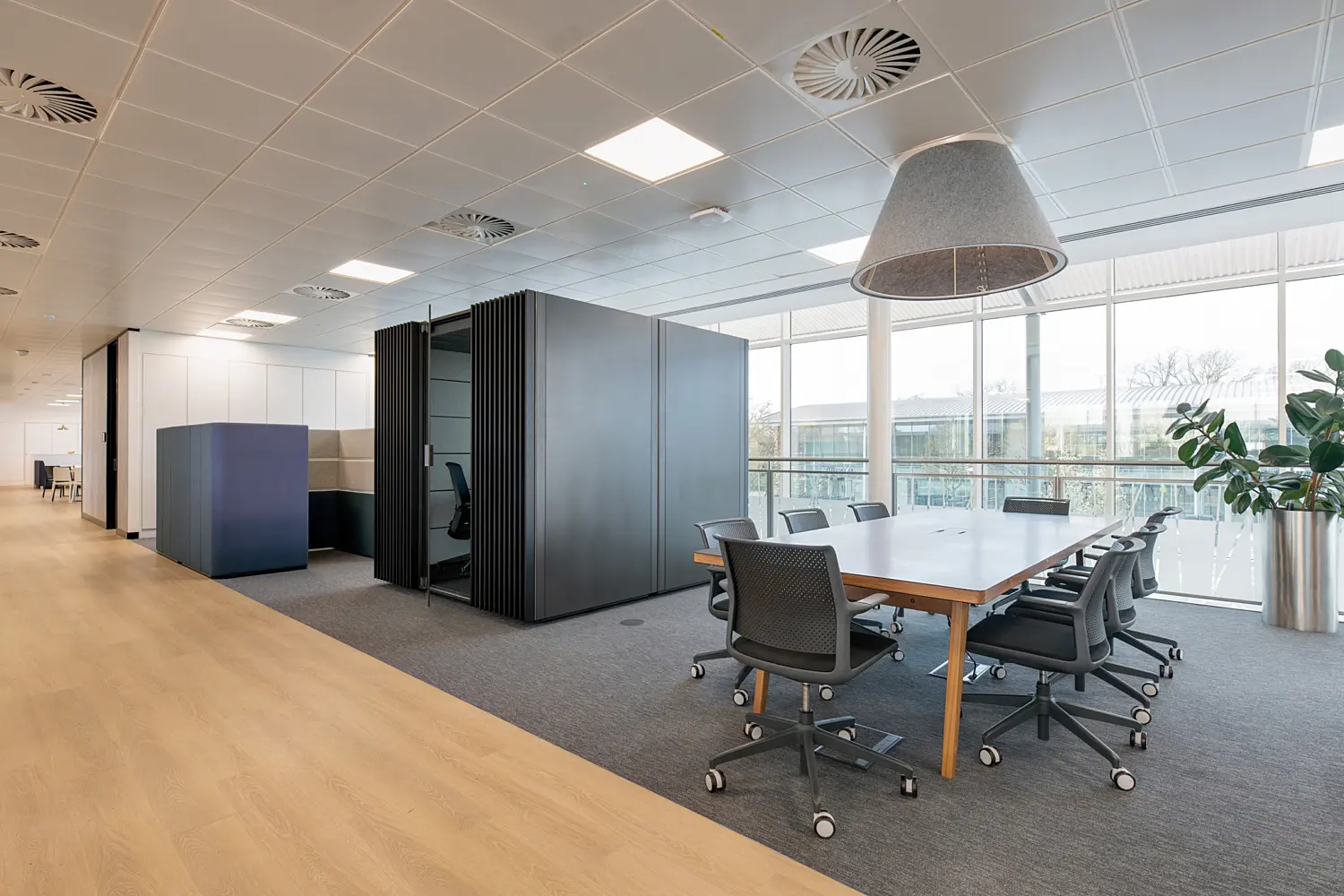 Modern open-plan office workspace featuring a large meeting table with task chairs positioned beside floor-to-ceiling windows. A grey acoustic pendant light hangs above the table, while a meeting pod and high-backed booth seating provide private spaces for focused work and conversations. Light wood flooring transitions to grey carpet within the collaboration area, and a large indoor plant adds greenery to the bright, contemporary environment. Natural daylight fills the space through extensive glazing overlooking neighbouring office buildings.