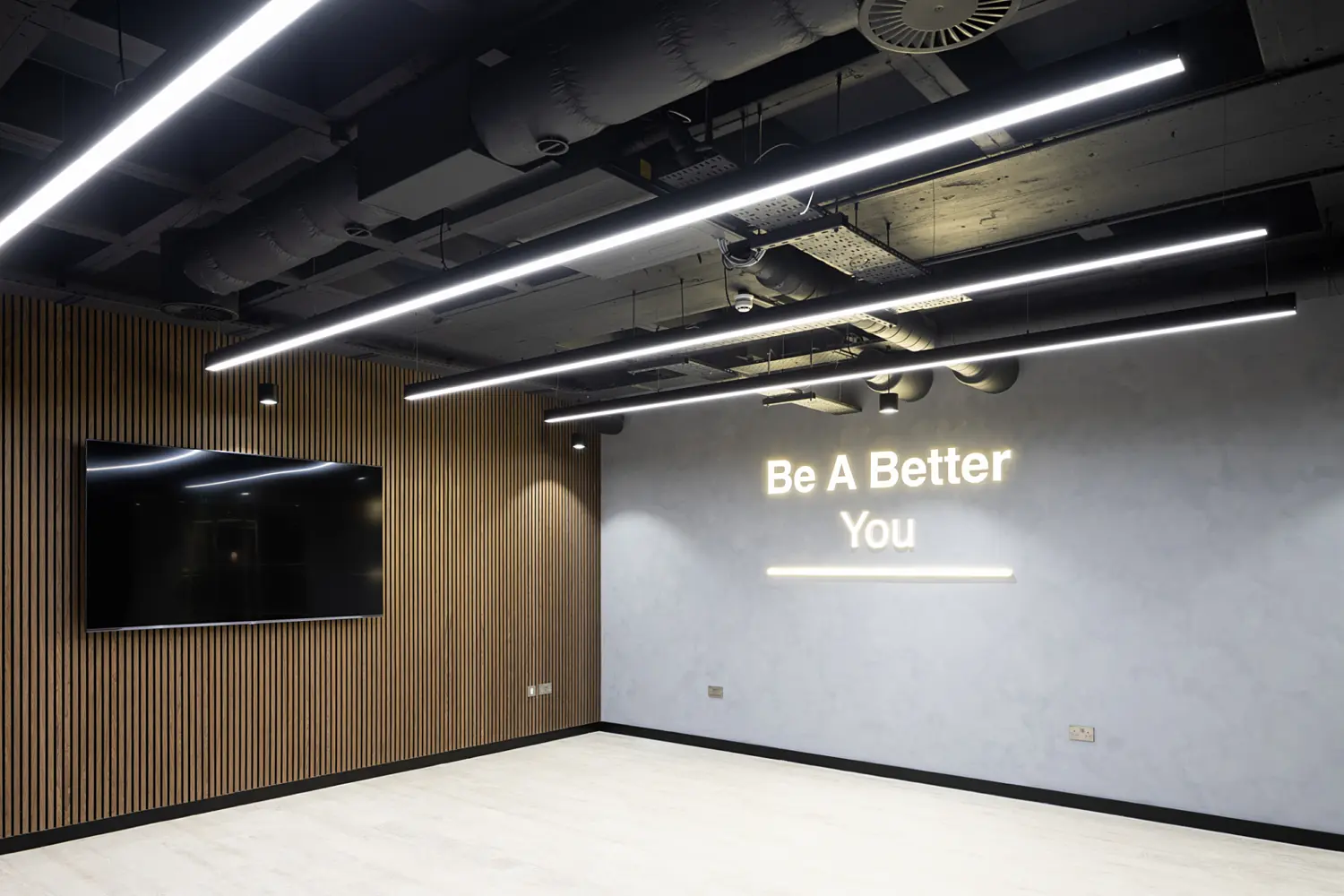 A contemporary office feature wall within a modern interior space. The main wall is finished in a smooth, light grey surface, displaying the illuminated text “Be A Better You” in soft white lettering, with a thin light bar beneath it. To the left, a large wall-mounted black screen sits against a contrasting vertical slatted wood panel. Above, the ceiling is open, revealing exposed ductwork, cables and services painted in dark tones, paired with several long, linear LED light fittings that run parallel across the space. The floor is a light wood-effect finish, keeping the area bright and uncluttered. The overall scene has a clean, minimal look, combining industrial ceiling elements with warm wood and soft, indirect lighting to create a bold focal point within the office.