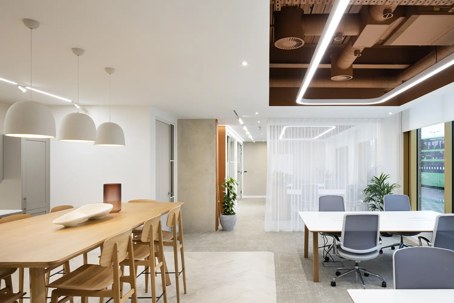 Wide view of a modern office interior that blends a breakout area with open workspaces. On the left, a long light wood high table is surrounded by matching wooden stools, with three large white pendant lights hanging above to create a soft, focused glow. The table sits on a light wood-effect floor, which transitions into a carpeted area on the right.  In the centre background, a sheer white curtain hangs from ceiling to floor, partially dividing spaces while still allowing light to pass through. Behind the curtain, additional seating and lighting are faintly visible. A corridor with neutral-toned walls and a door extends beyond this area, adding depth to the layout.  On the right, several white desks with grey office chairs are arranged near large windows that bring in natural daylight and offer a view of a bus and street outside. A potted green plant is placed near the window, adding a natural element.  Above, the ceiling combines a clean white finish with a feature section of exposed services painted in a warm brown tone, framed by continuous LED strip lighting. The overall palette of soft whites, warm wood and greenery creates a bright, calm and contemporary workplace environment.